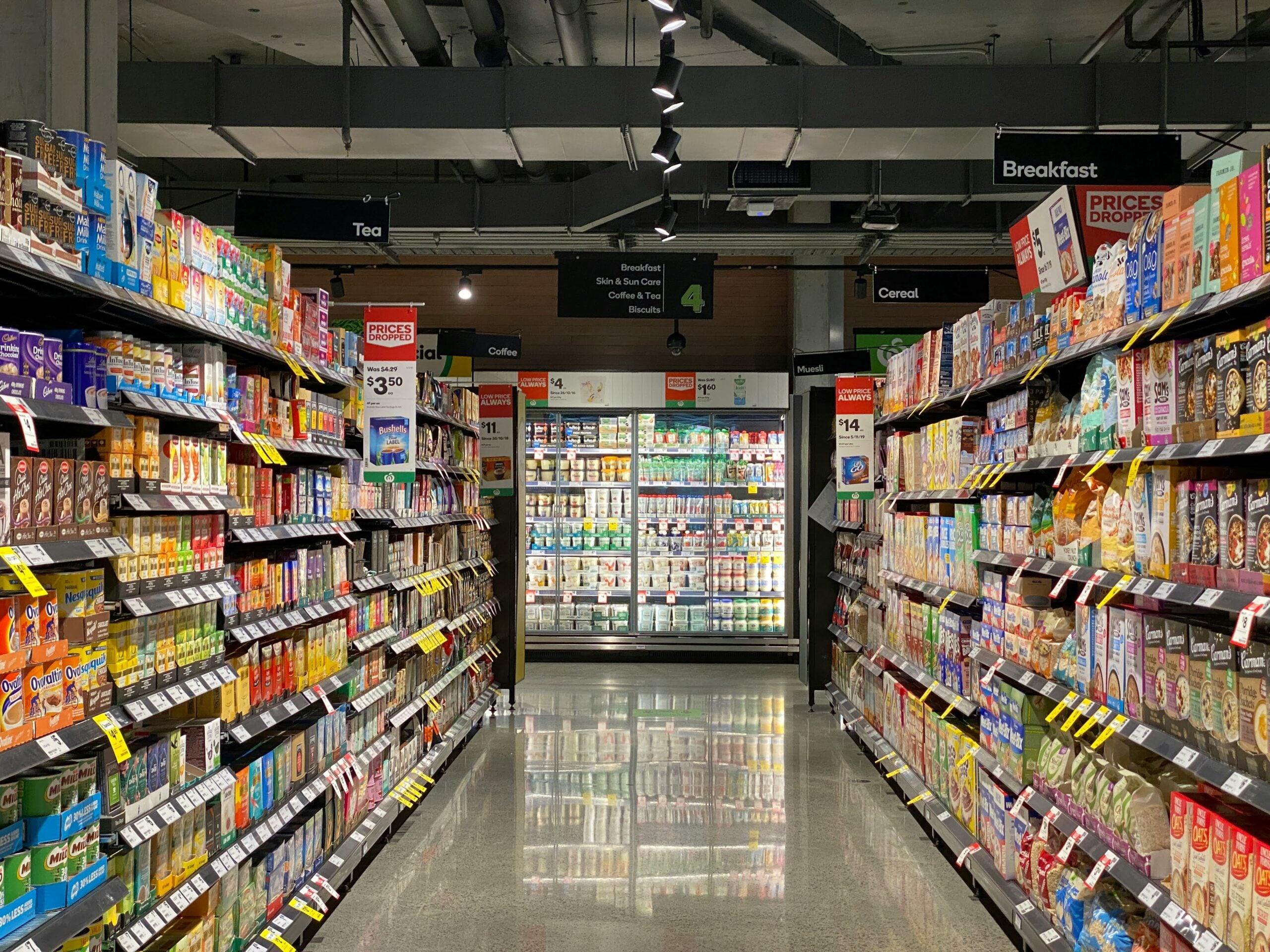 A supermarket aisle stocked with various boxed and packaged food items, mainly cereals. Signs above point to sections for tea, coffee, breakfast, and cereal. Refrigerated dairy products are visible at the end of the aisle.