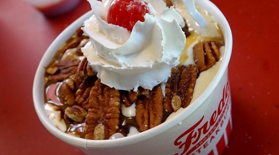 A cup of ice cream topped with whipped cream, a cherry, pecan halves, and caramel sauce, served in a white and red-striped container on a red surface.