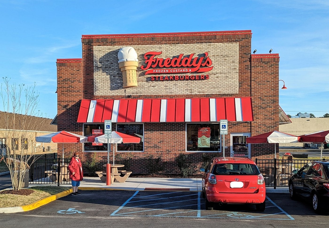 A brick Freddy’s Frozen Custard & Steakburgers restaurant with a large ice cream cone sign, red-striped awning, outdoor seating, and two parked cars in front, one next to a person in a red coat.