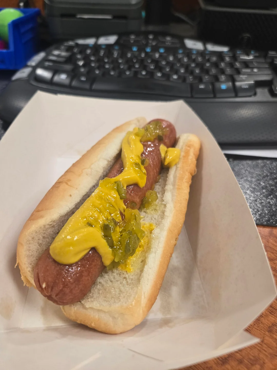 A Fast-Food Hot Dog in a bun topped with mustard and relish sits in a paper tray on a desk, with a computer keyboard and office supplies visible in the background.
