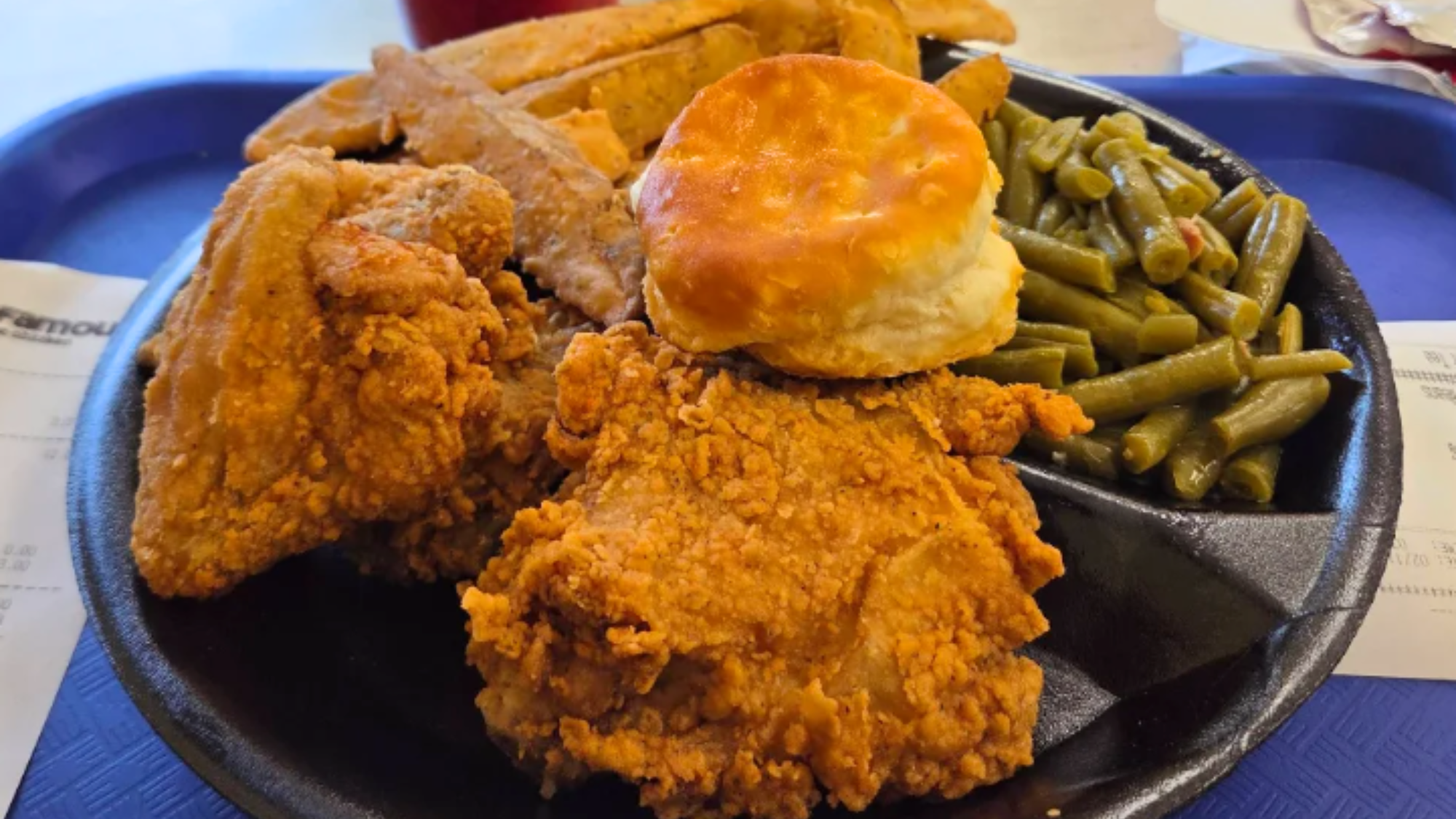 A classic meal from popular fried chicken chains: crispy chicken pieces, a biscuit, green beans, and thick-cut fries served on a black tray with receipts visible underneath.