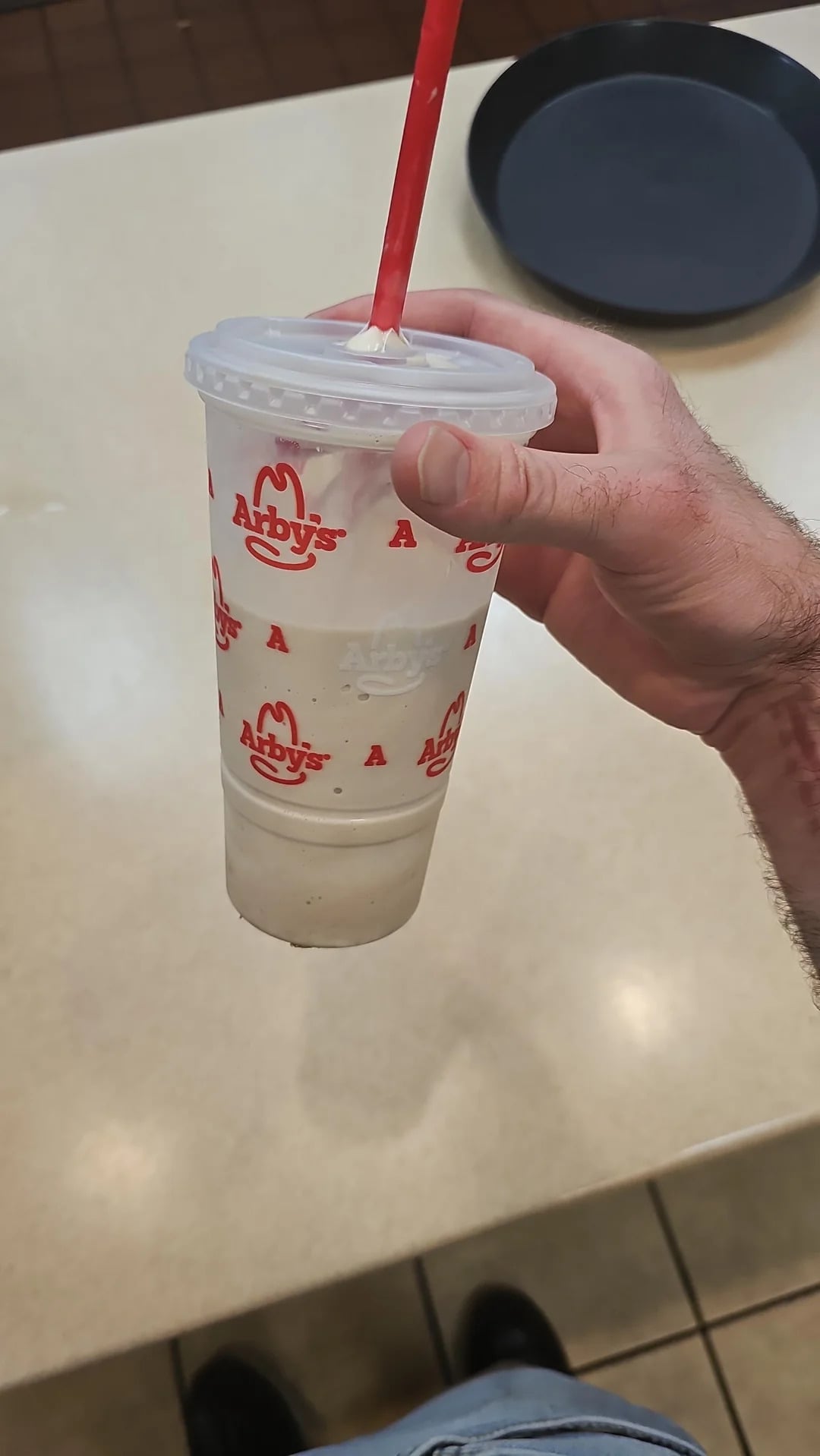 A hand holds a nearly empty Arby's cup with a red straw, featuring the Arby's logo and branding. The cup appears to contain remnants of one of Arby's popular fast-food desserts, such as a milkshake or soft drink.
