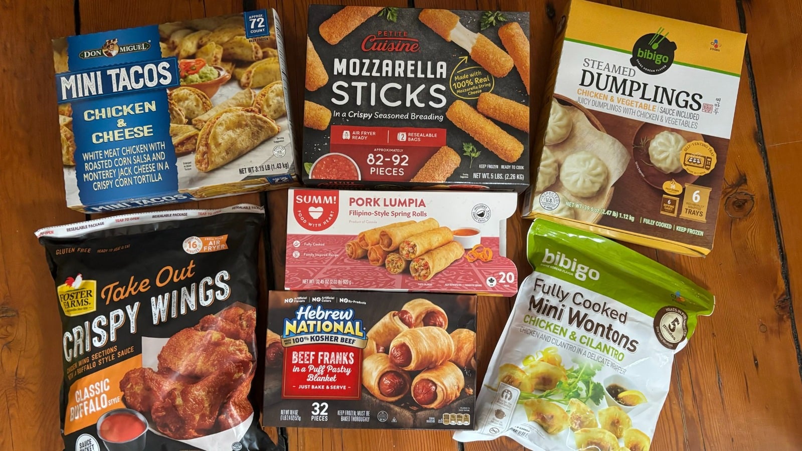 Assorted boxes and bags of frozen appetizers and snacks, including mini tacos, mozzarella sticks, steamed dumplings, chicken wings, pork lumpia, beef franks, and mini wontons, arranged on a wooden table.