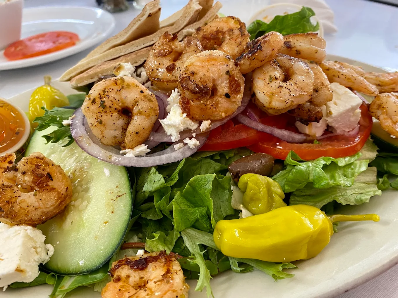 A Greek salad topped with grilled shrimp, feta cheese, cucumber, tomato, red onion, pepperoncini, and pita bread on a white plate, with salad dressing and sauce in small containers nearby.