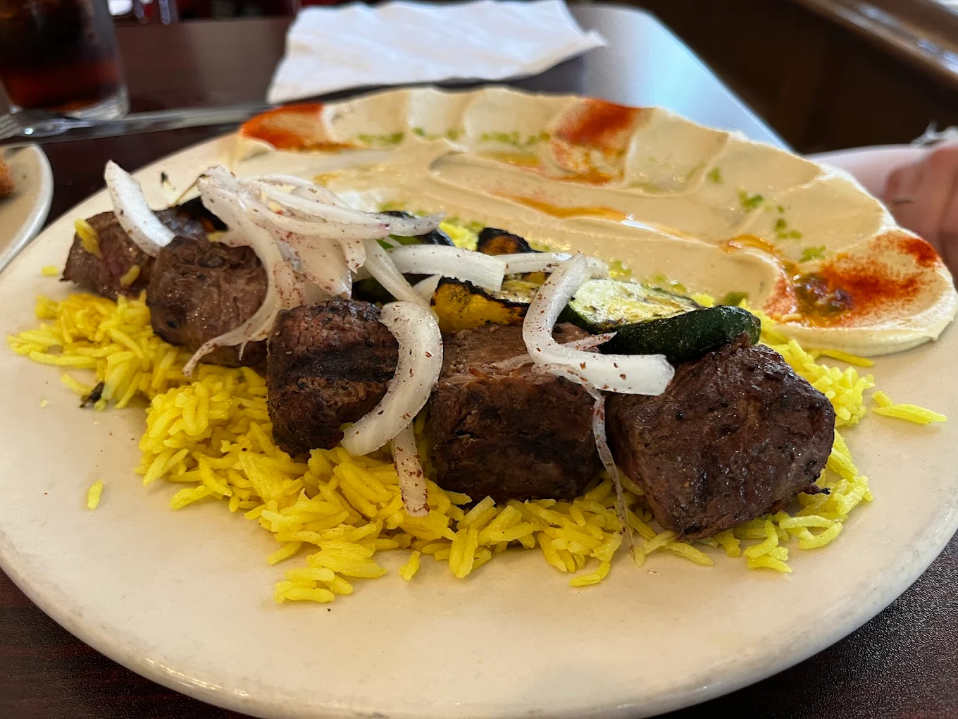 A plate with yellow rice, grilled beef kebabs, sliced onions, grilled zucchini, and a spread of hummus garnished with paprika and oil on the side.