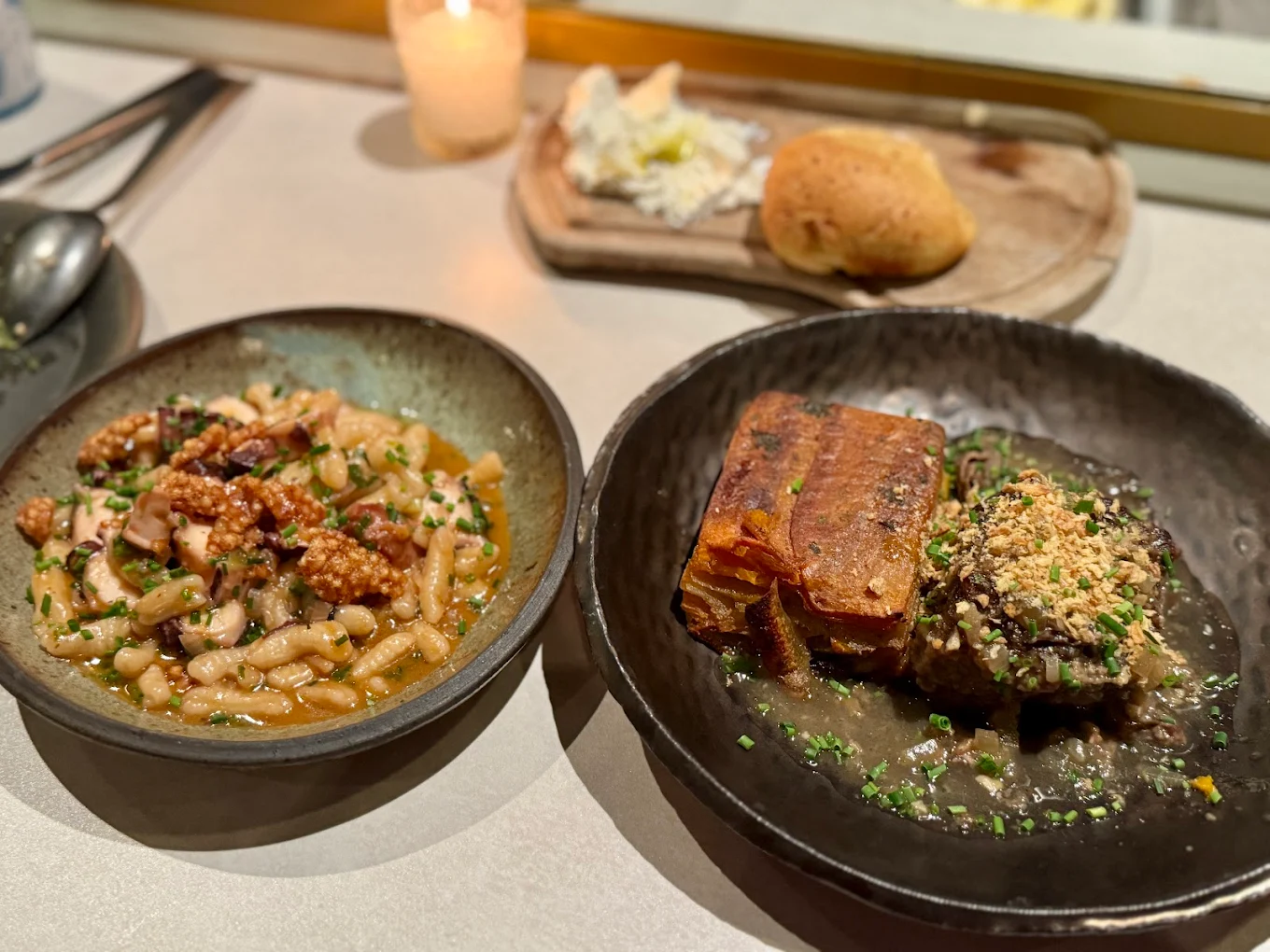 Two plates of gourmet food on a table: one with pasta and what appears to be octopus, the other with braised meat topped with breadcrumbs and a crispy rectangular garnish. Bread and cheese are in the background. A candle is lit nearby.