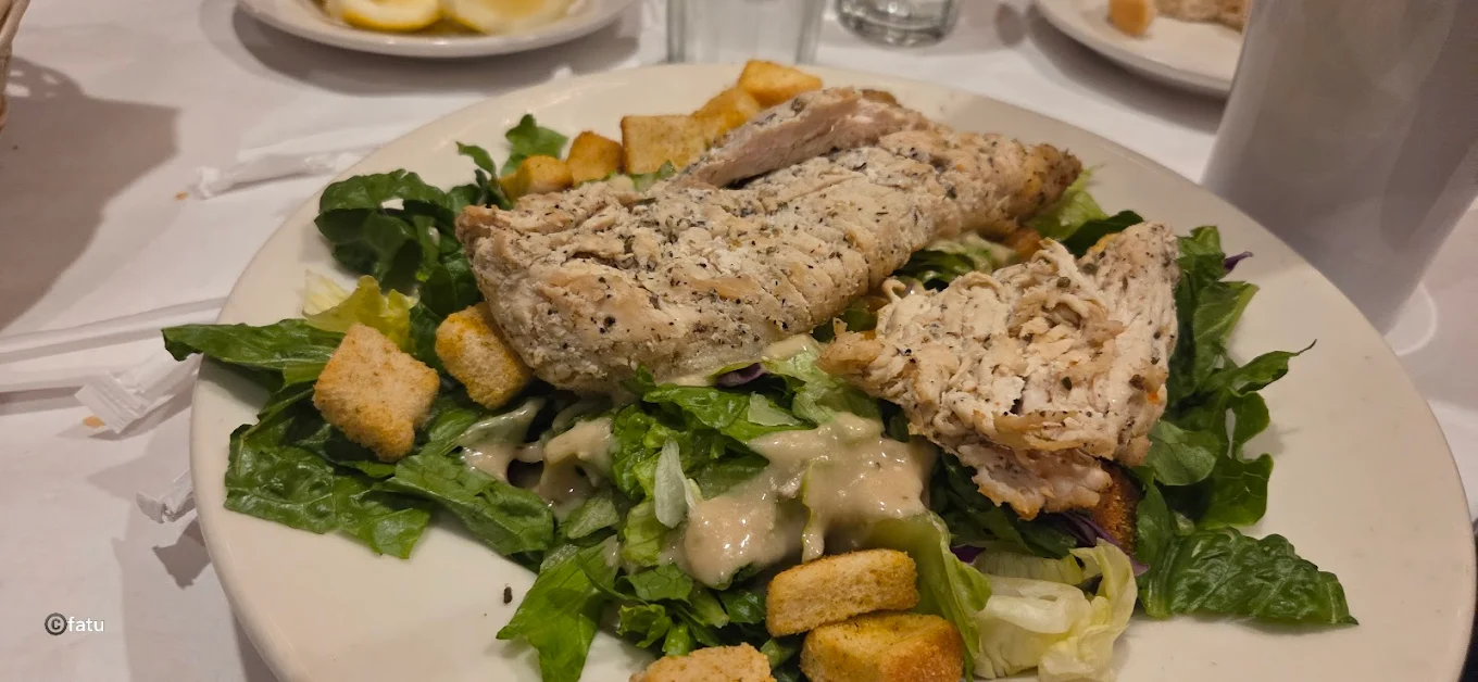 A plate of salad topped with grilled chicken breast, croutons, and creamy dressing, served on a bed of leafy greens; drinks and lemon slices are visible in the background.