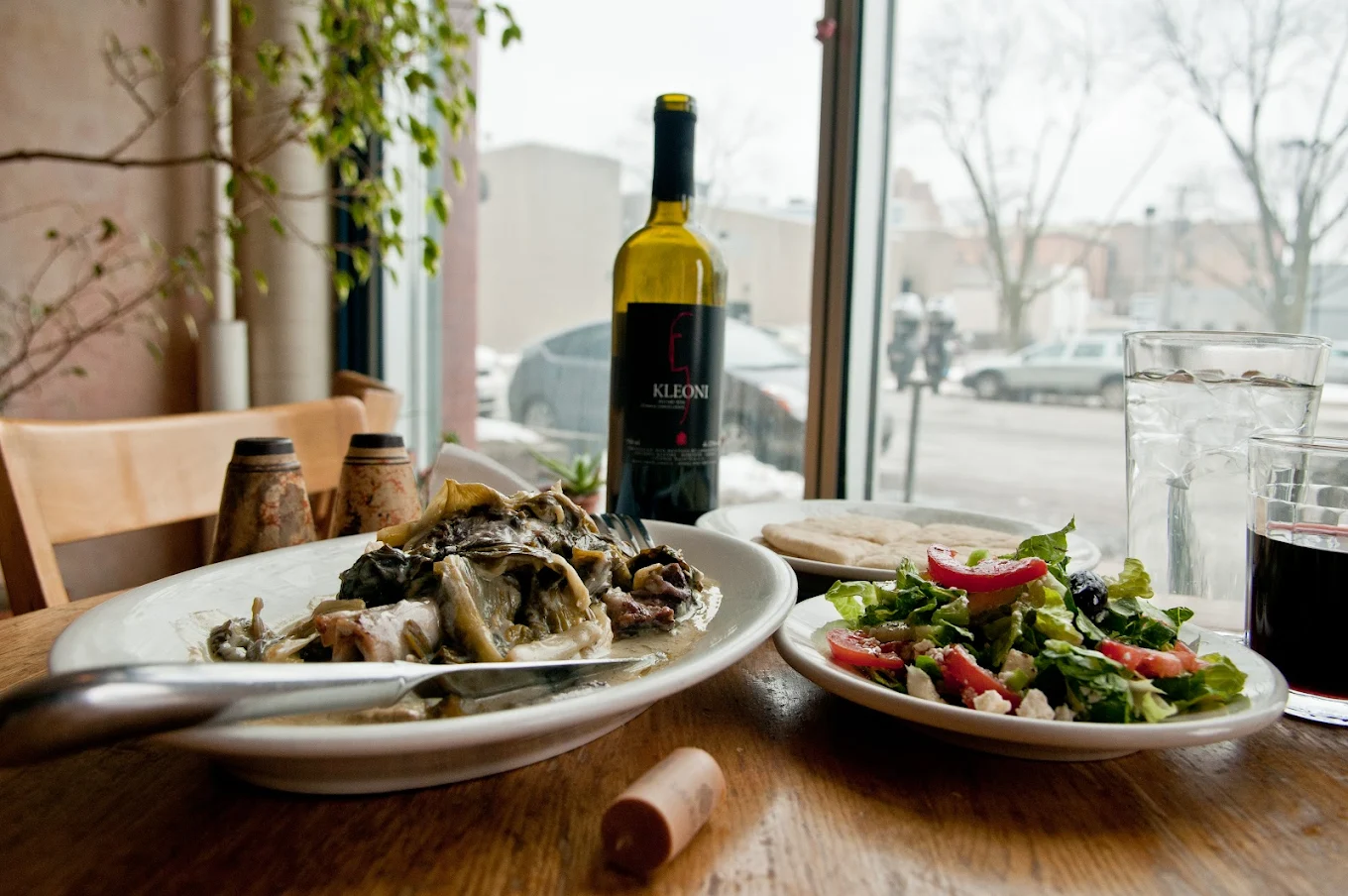 A table by a window holds a plate of cooked fish, a side salad with tomatoes and olives, a wine bottle, a wine glass, water glasses, and flatbread. Snowy street and trees are visible outside.