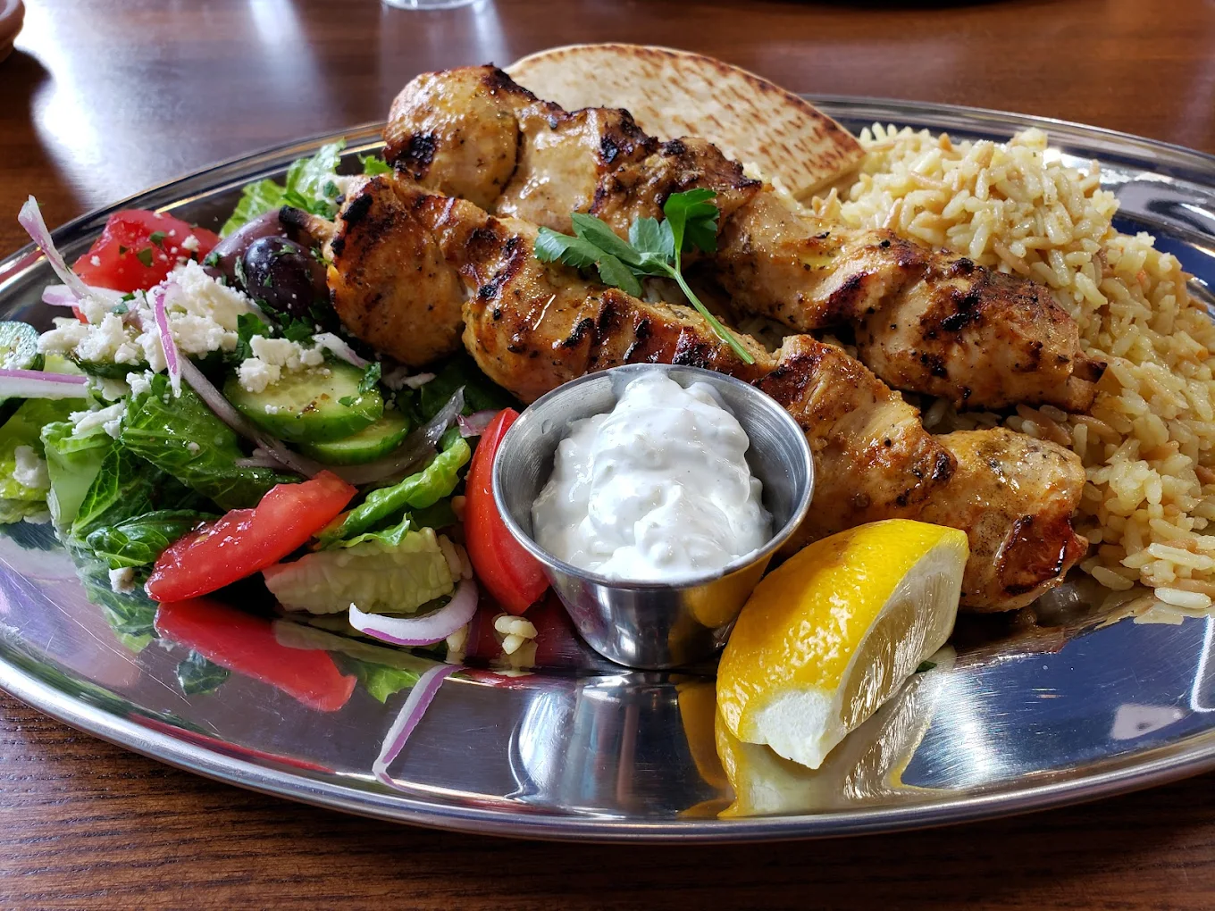 A metal plate with grilled chicken skewers, yellow rice, a Greek salad with tomatoes, cucumber, olives, and feta, a slice of pita bread, a lemon wedge, and a small cup of tzatziki sauce.