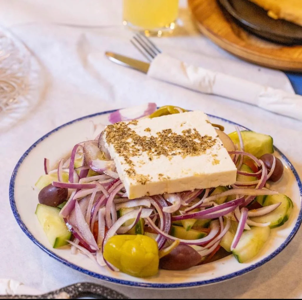 A plate of Greek salad with sliced cucumbers, tomatoes, red onions, olives, and peppers, topped with a thick slice of feta cheese sprinkled with herbs. A fork and knife wrapped in a napkin are in the background.