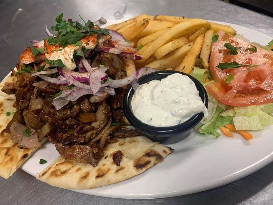 A plate with pita bread topped with seasoned meat, sliced onions, herbs, and paprika, served with French fries, a cup of tzatziki sauce, and a side salad of lettuce and tomatoes.