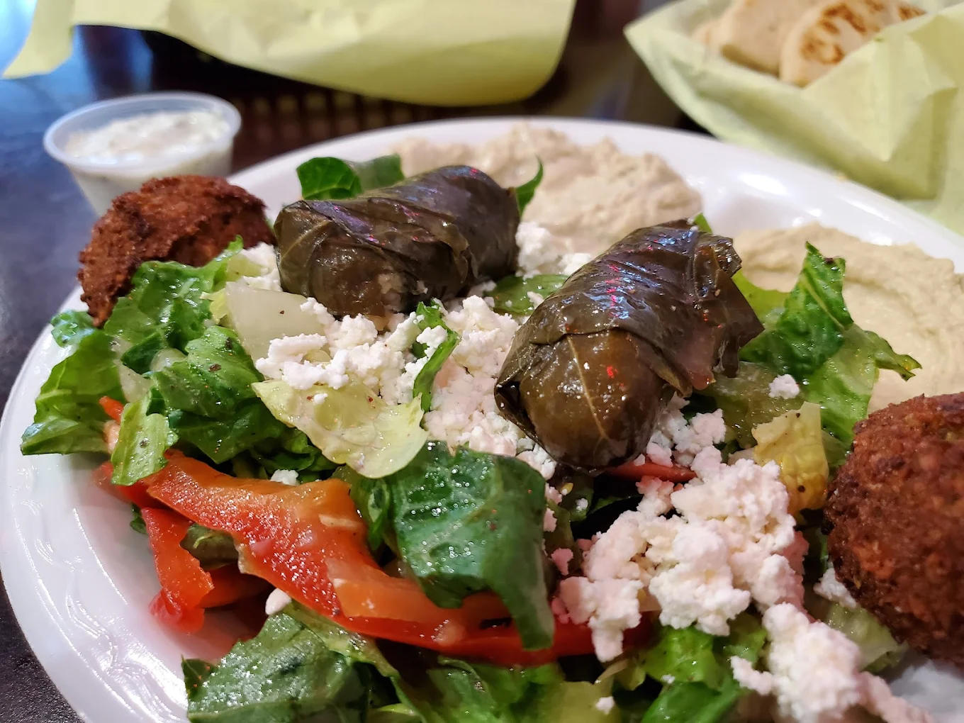A plate of Mediterranean food with grape leaves, falafel, salad topped with feta cheese, red bell peppers, and hummus, with a side of sauce and pita bread in the background.