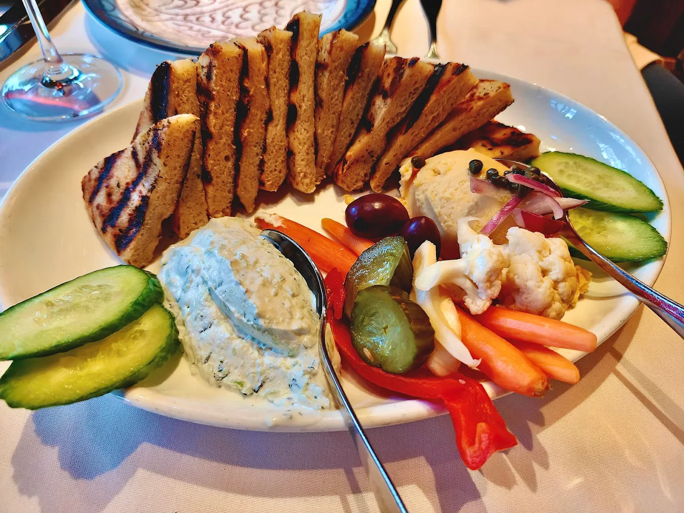 A plate with grilled bread slices, cucumber, olives, pickles, red bell pepper, carrot sticks, pickled cauliflower, and two dips, served on a white tablecloth.