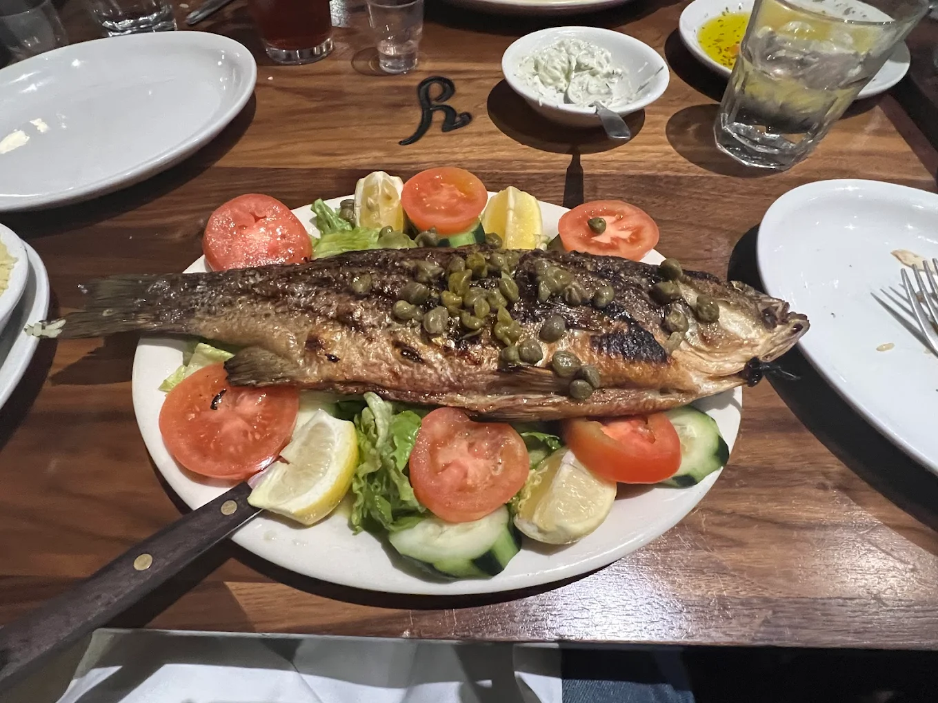 A whole grilled fish topped with capers, served on a plate with sliced tomatoes, cucumbers, lettuce, and lemon wedges. The plate is on a wooden table with drinks and small bowls nearby.