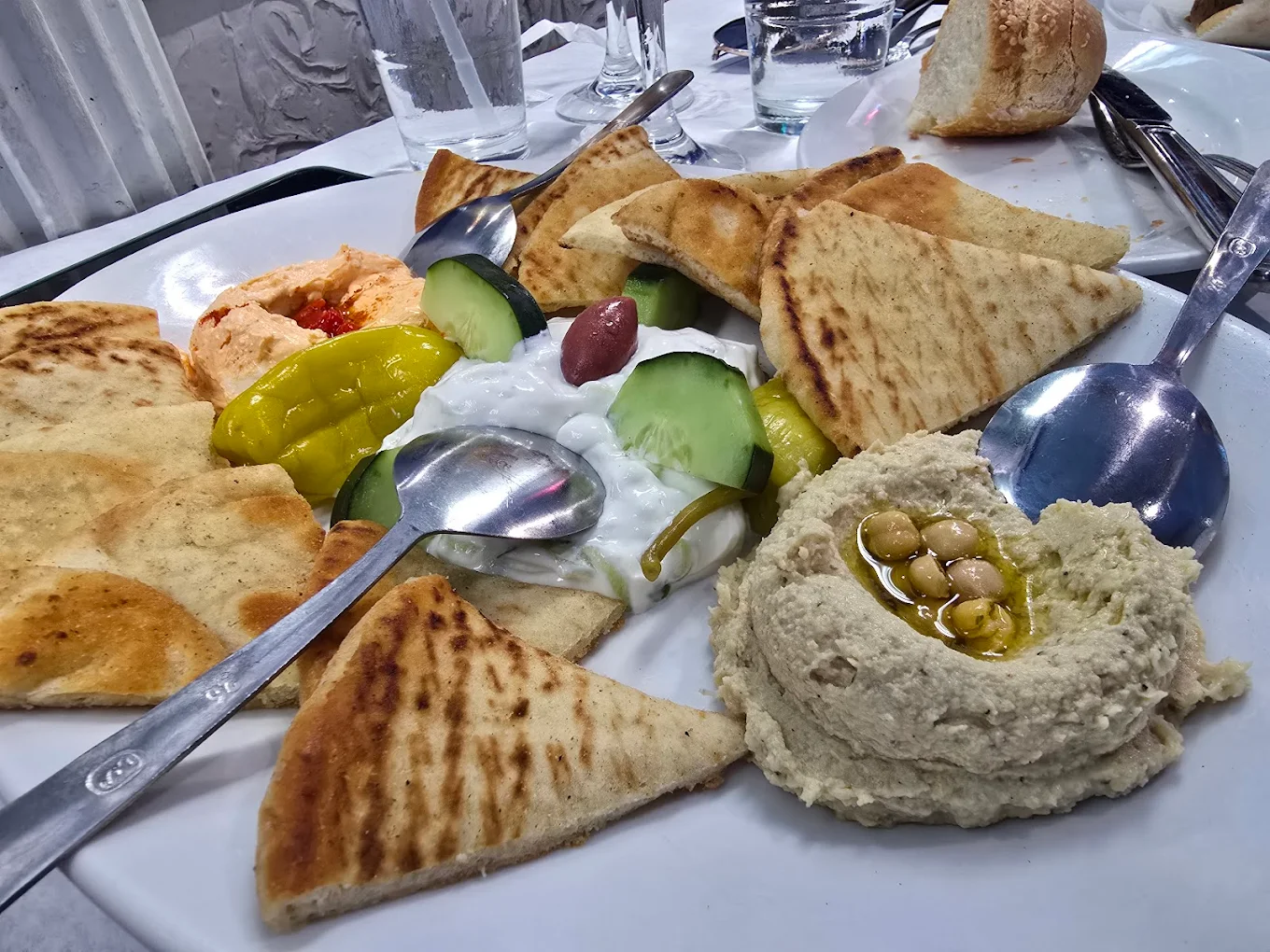 A plate with sliced pita bread, hummus topped with chickpeas and olive oil, tzatziki with cucumber and olives, and a portion of red pepper dip, served with metal spoons and a piece of bread in the background.