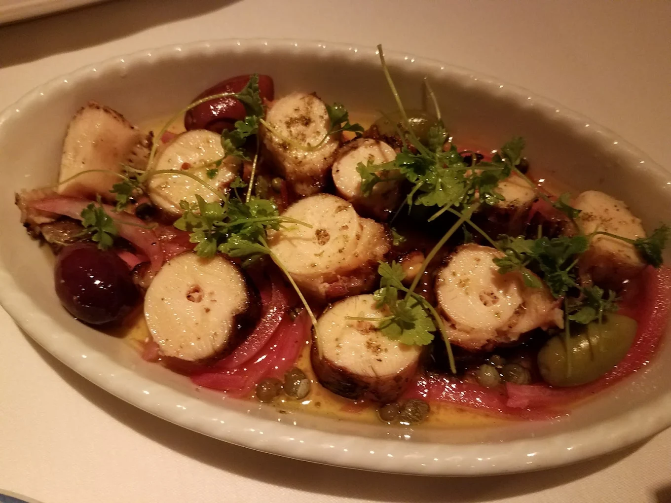 Sliced grilled octopus served on a white oval plate with olives, capers, pickled onions, sauce, and garnished with fresh herbs. Best Greek restaurants in each state