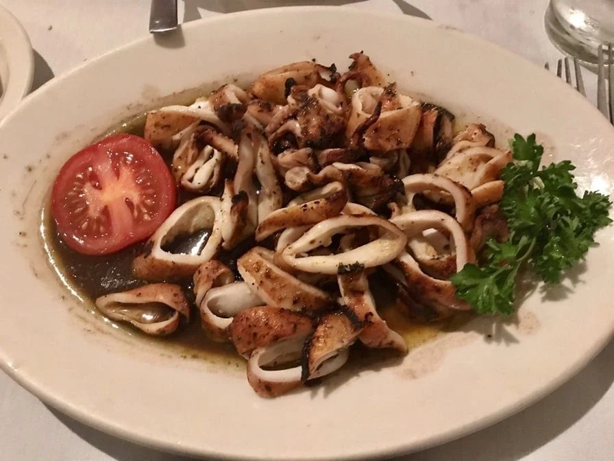 A white plate with grilled calamari rings in sauce, garnished with a slice of tomato and a sprig of parsley. A fork, knife, and part of another plate are visible in the background.