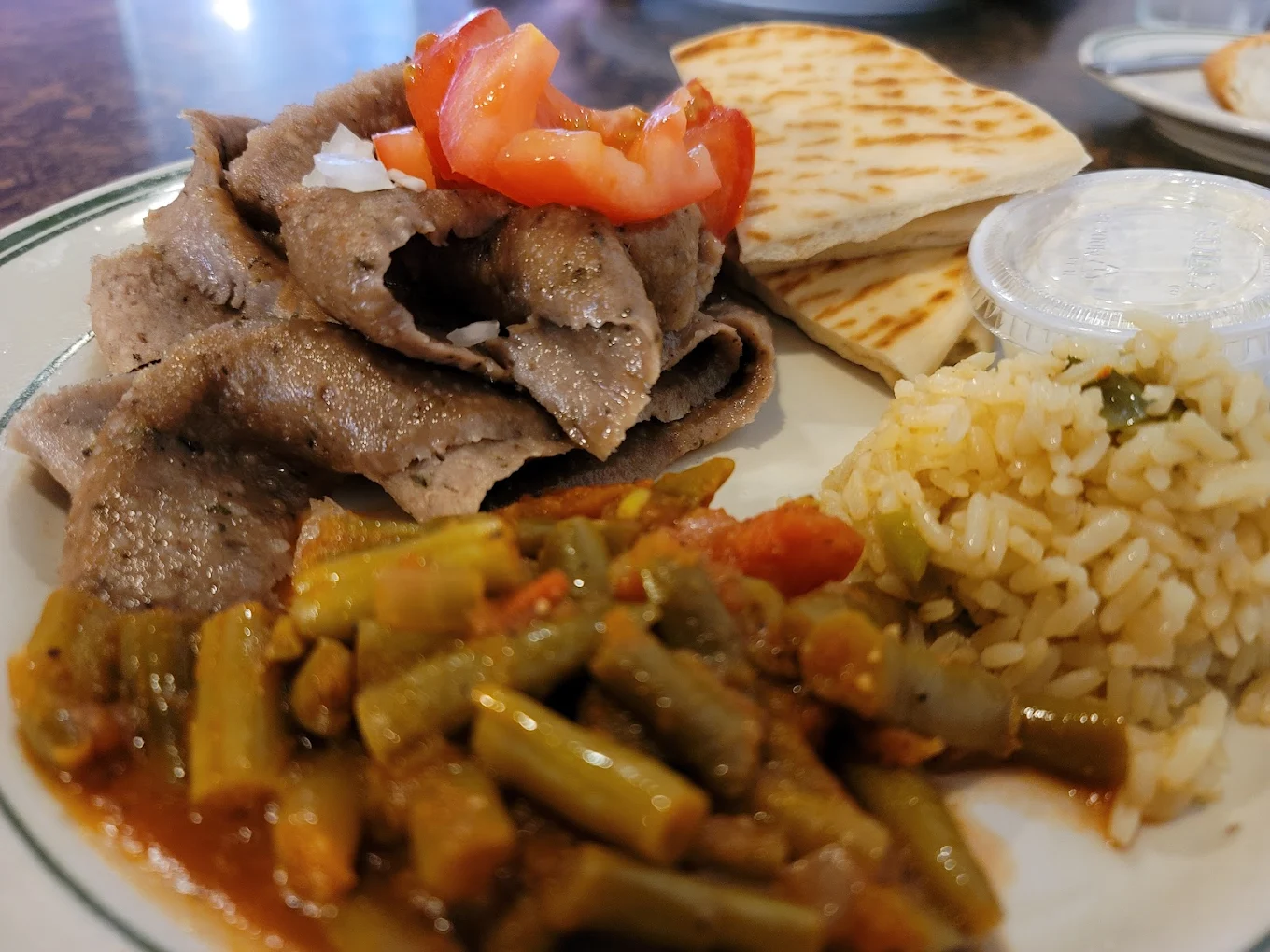 A plate with gyro meat topped with onions and tomatoes, pita bread, a serving of yellow rice, green beans in tomato sauce, and a container of white sauce.