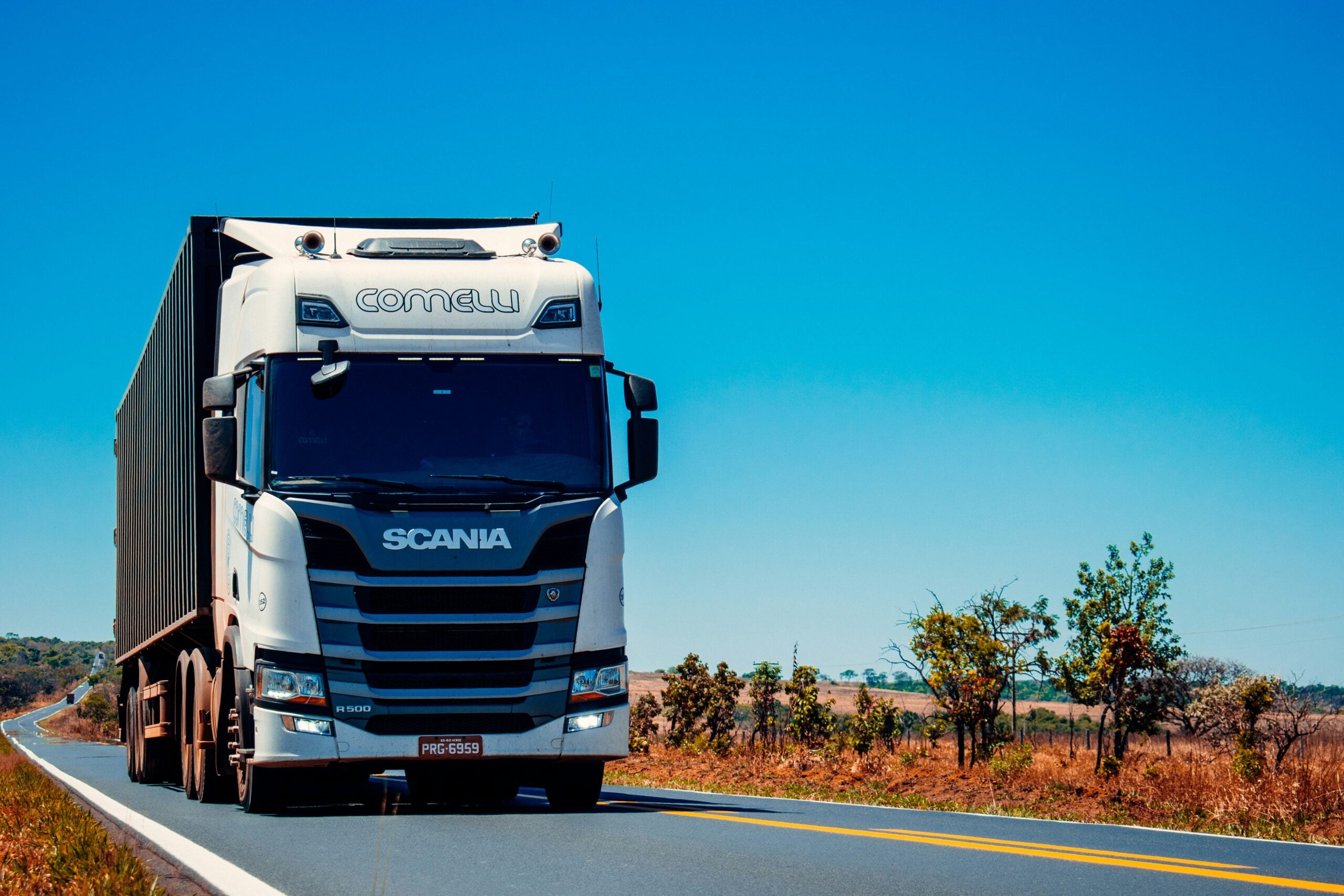 A white Scania semi-truck with a large trailer drives along a rural highway surrounded by dry grass and scattered trees under a clear blue sky.