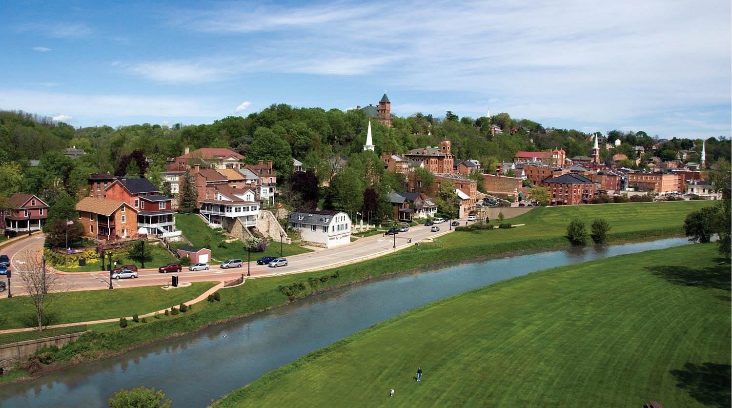 A scenic view of one of the unique American towns, with historic brick buildings and churches on a hillside, a winding river in the foreground, and green trees and grass under a partly cloudy sky.
