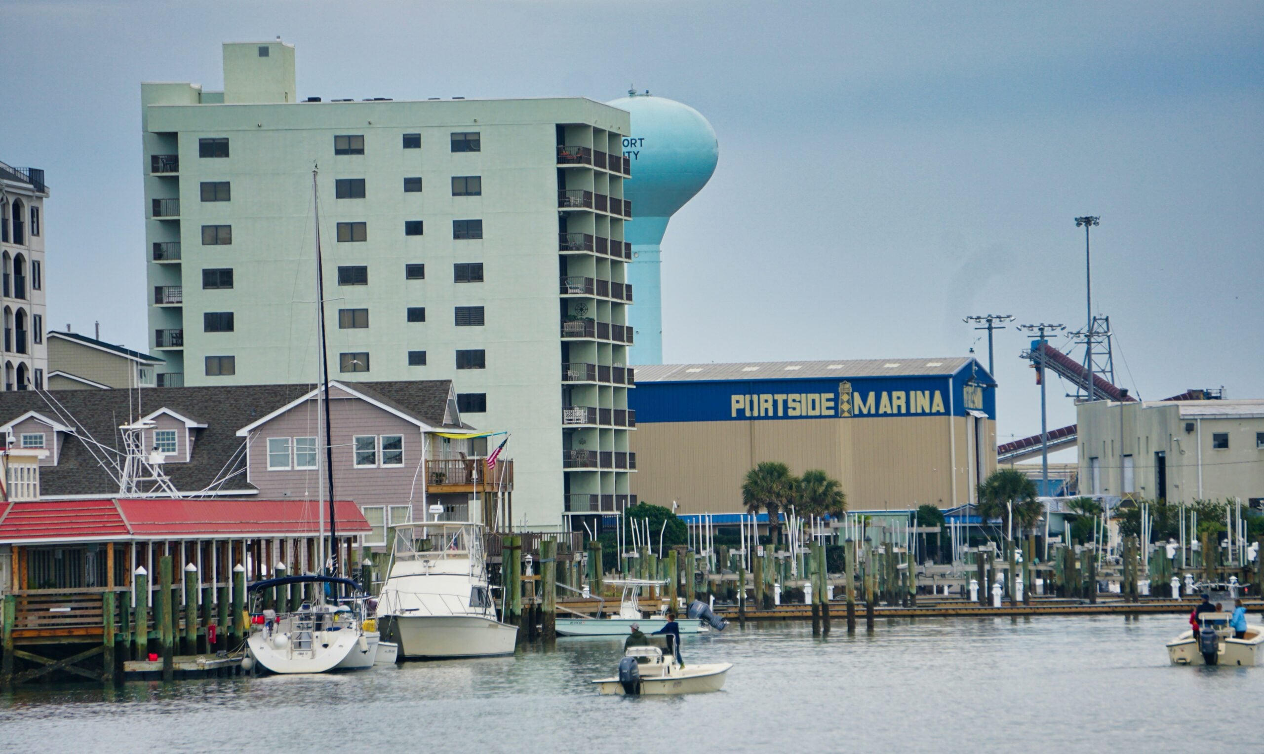 Boats docked at a marina with people on the water, surrounded by buildings, including a multi-story hotel and a large blue water tower labeled "PORTSIDE MARINA." The sky is overcast.