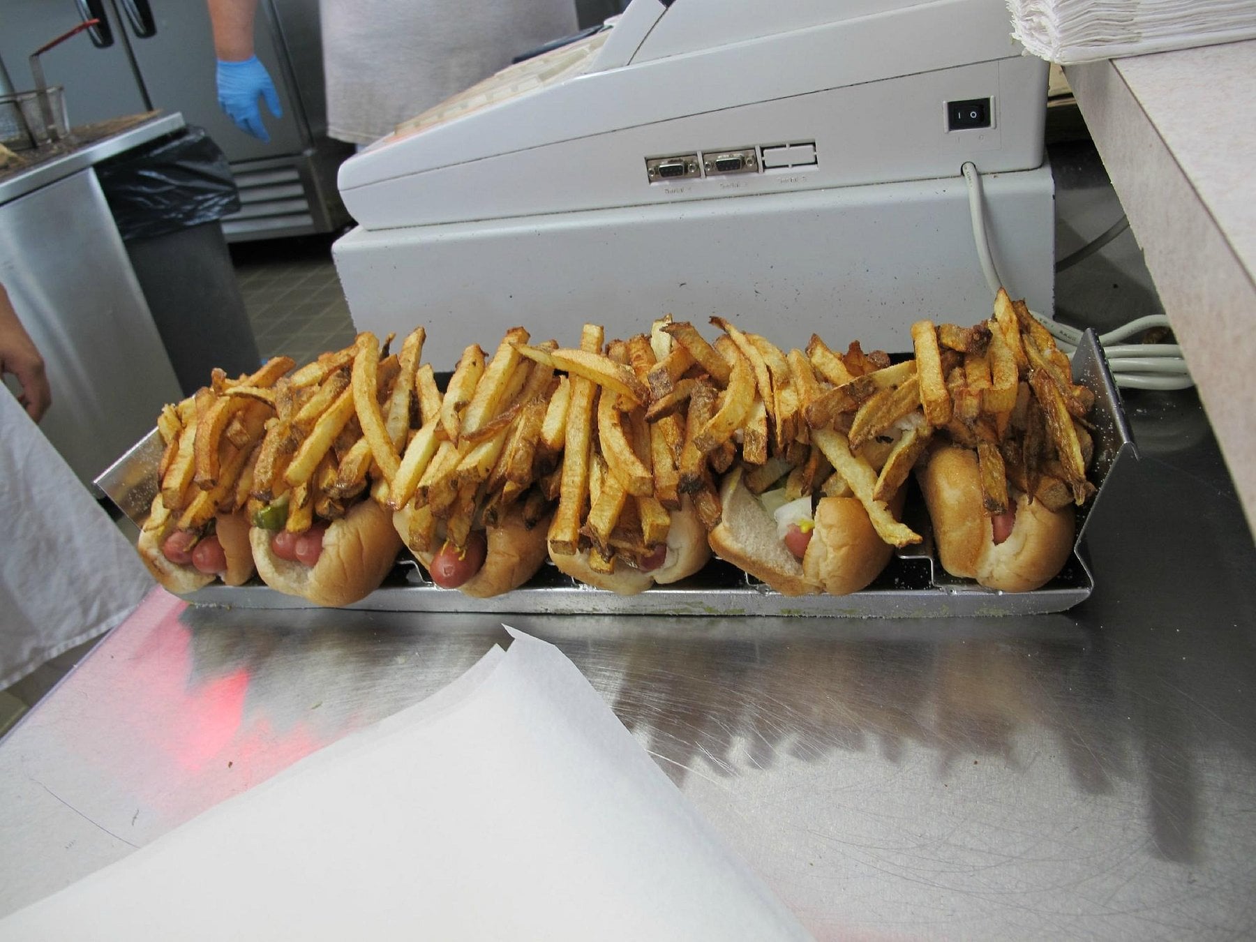 A metal tray holds several hot dogs piled high with French fries, sitting on a counter next to a cash register—classic road trip food stops fare in a bustling restaurant kitchen.