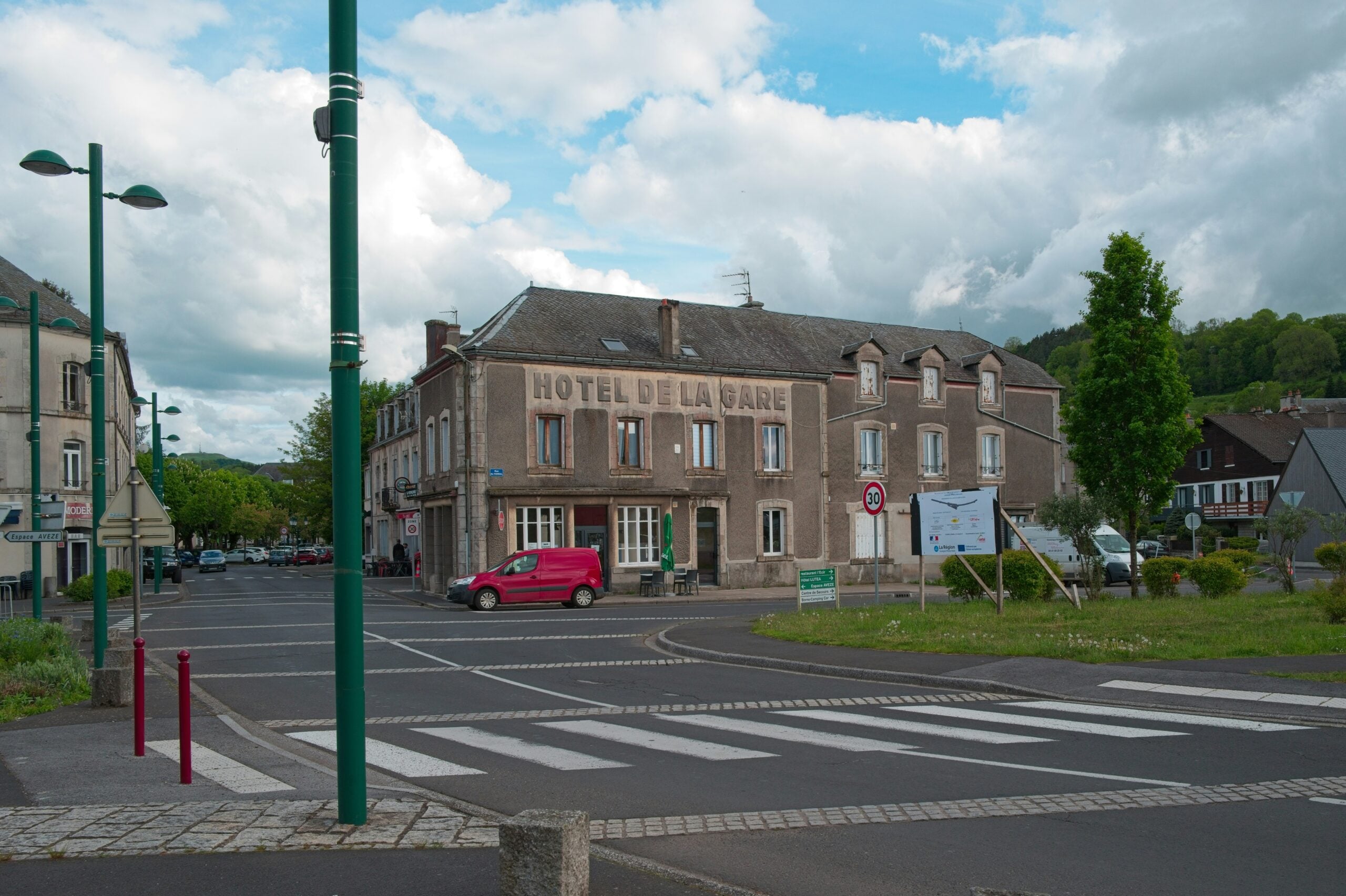A street view of a stone building labeled "HOTEL DE LA GARE" with a red van parked in front, crosswalks, street lamps, and a partly cloudy sky in a small town setting.