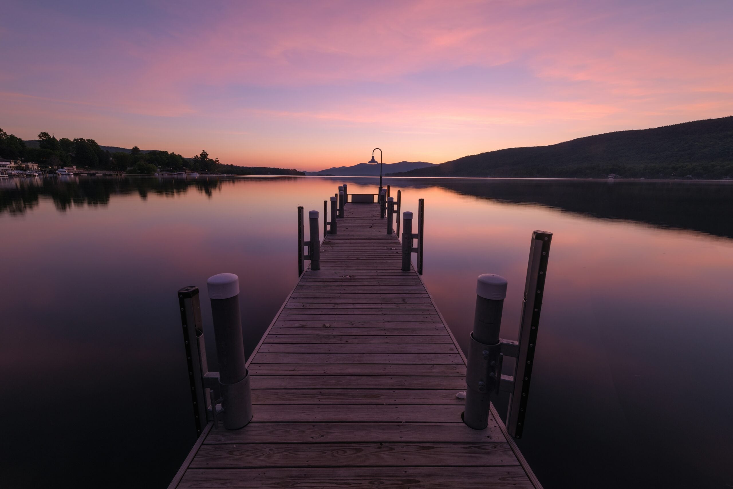 A wooden dock extends over a calm lake at sunset, with soft pink and purple hues in the sky, mountains in the background, and reflections of the colorful sky on the still water.