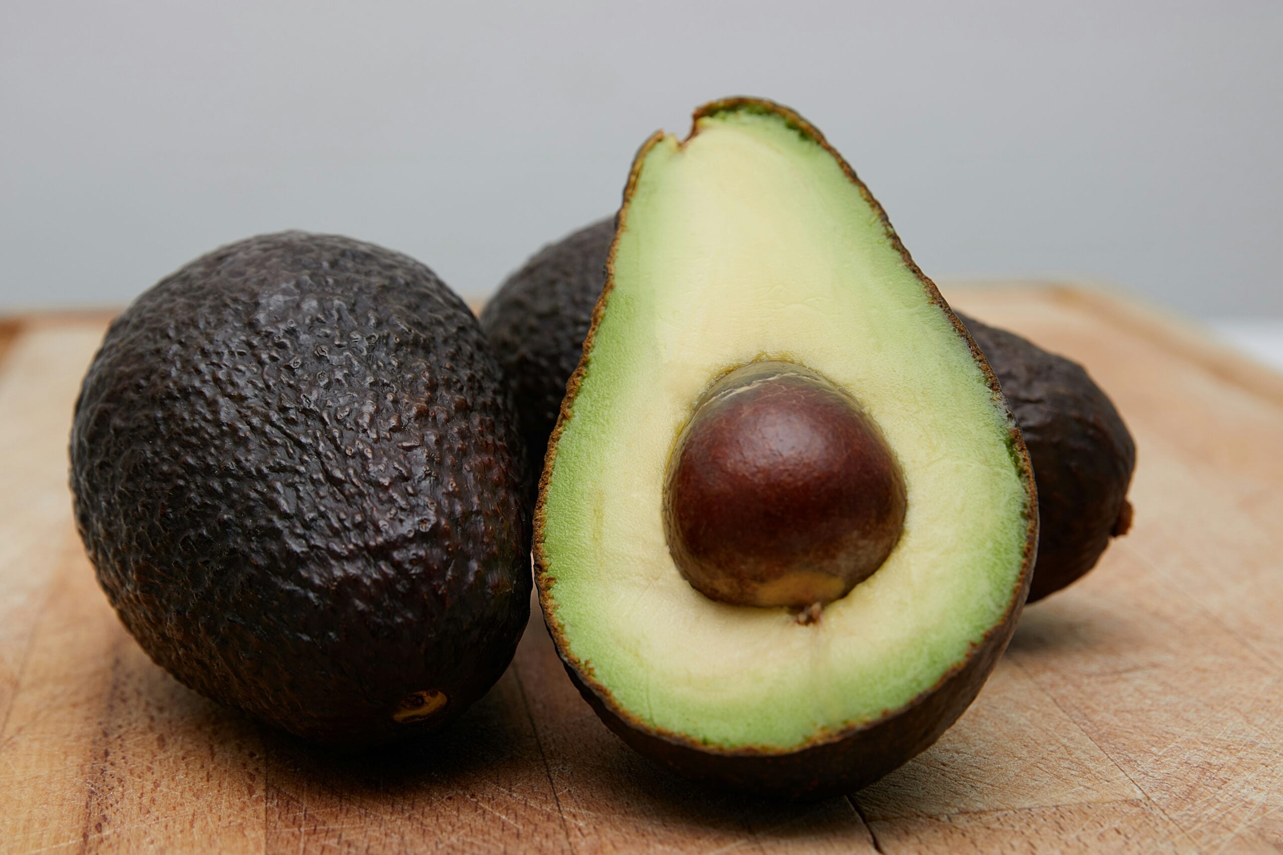 Three avocados on a wooden surface, with one cut in half showing its green flesh and large brown seed, while the other two remain whole with dark, textured skin.
