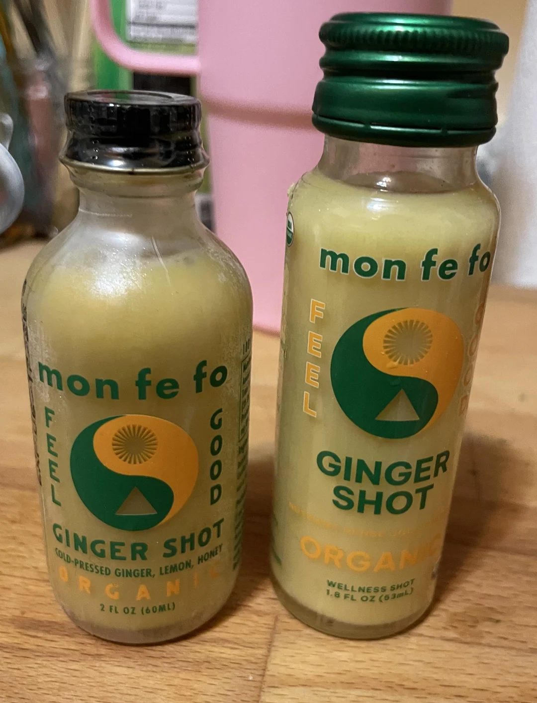 Two bottles of "mon fe fo" organic ginger shots with green caps and labels, containing ginger, lemon, and honey. They are placed on a wooden surface with a pink object in the background.