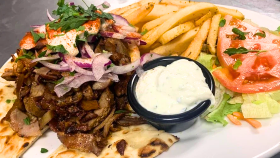 A plate with seasoned grilled meat on flatbread, topped with sliced red onions and herbs, served with thick-cut fries, creamy white sauce, and a fresh salad&mdash;just like you&rsquo;d find at the best Greek restaurants in each state.