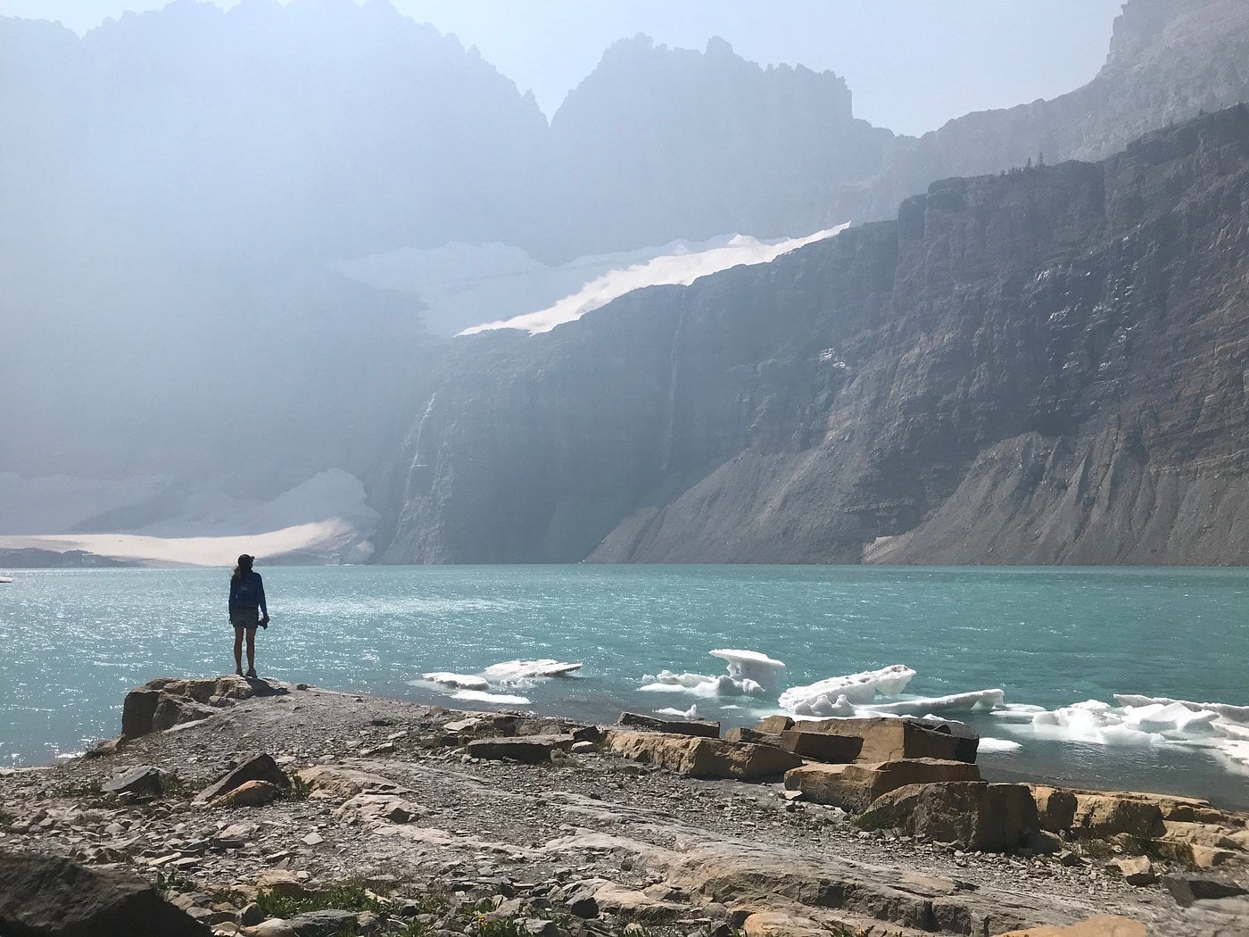 A person stands on a rocky shoreline, gazing at a turquoise glacial lake with floating ice, surrounded by steep, misty mountains—evoking the breathtaking scenery found near some Unique American Towns.