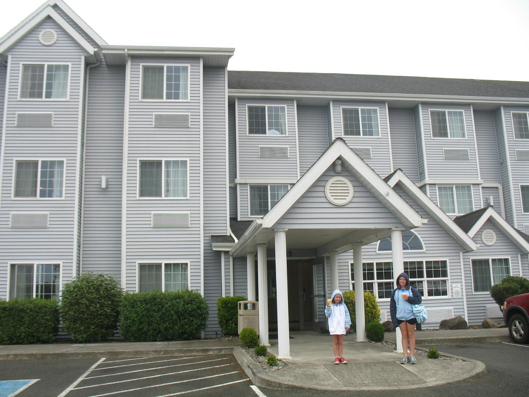 Two people in hooded jackets stand under the entrance of a gray, three-story hotel building with white trim. Shrubs line the property, and an overcast sky adds to the scene—a glimpse into what Hotel Brands 2026 might look like.
