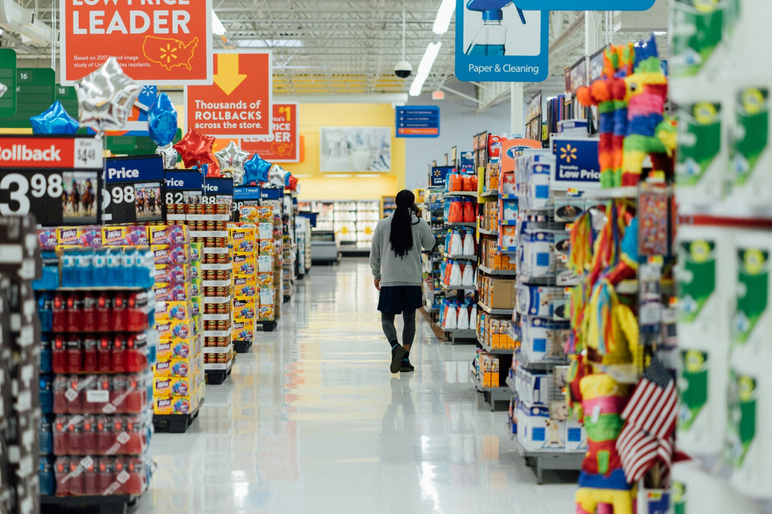 A person walks down a brightly lit grocery store aisle lined with various products, colorful signs, and balloons. Price tags and promotional banners hang from the ceiling, and shelves are fully stocked.