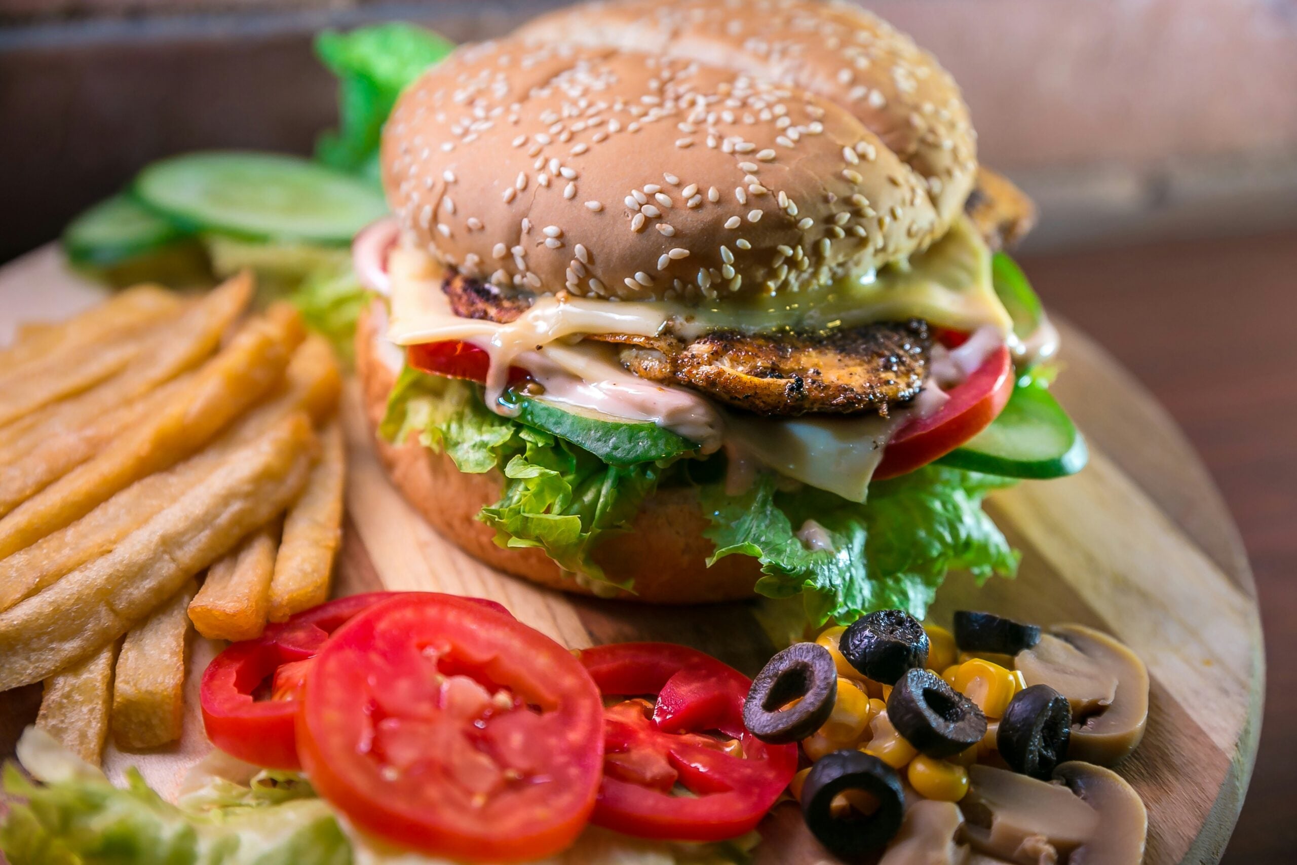 A sesame seed burger with lettuce, tomato, cheese, and grilled patty sits on a wooden plate, accompanied by French fries, sliced tomatoes, cucumber, olives, corn, and mushrooms.