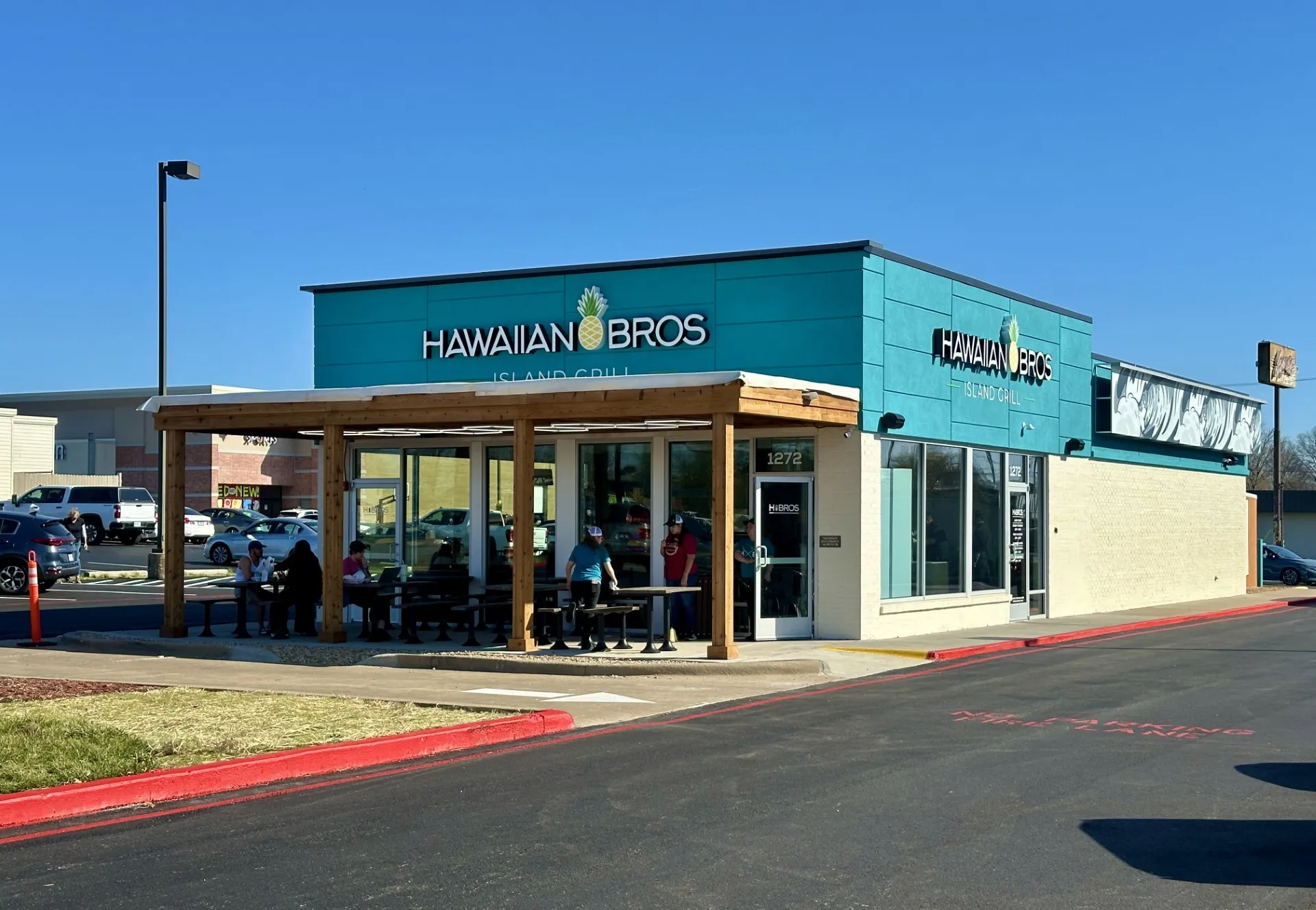 A Hawaiian Bros Island Grill restaurant with teal and white exterior, large windows, and a wooden patio where people are sitting and eating under a clear blue sky. Cars are parked nearby.