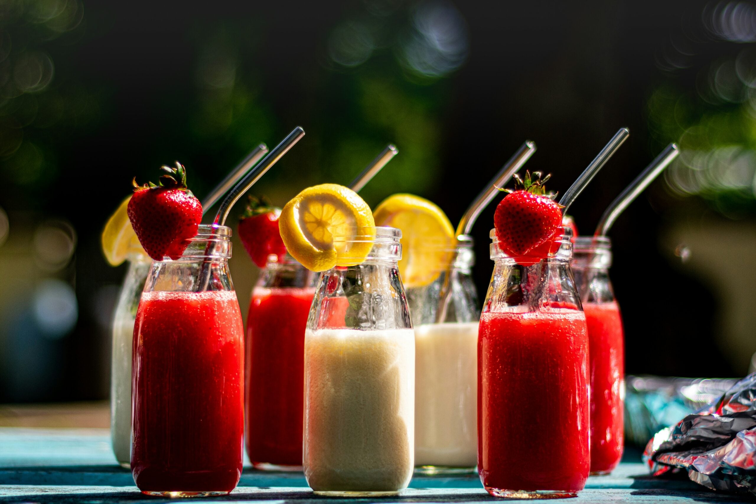 Six glass bottles with metal straws are filled with red and white drinks, garnished with fresh strawberries and lemon slices, sitting on a blue table outdoors with sunlight and a blurred green background.