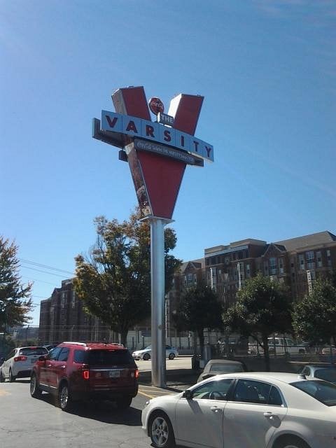 A large red and white “V” sign for The Varsity stands tall in a parking lot, a classic landmark among road trip food stops, surrounded by parked cars and trees, with buildings in the background under a clear blue sky.