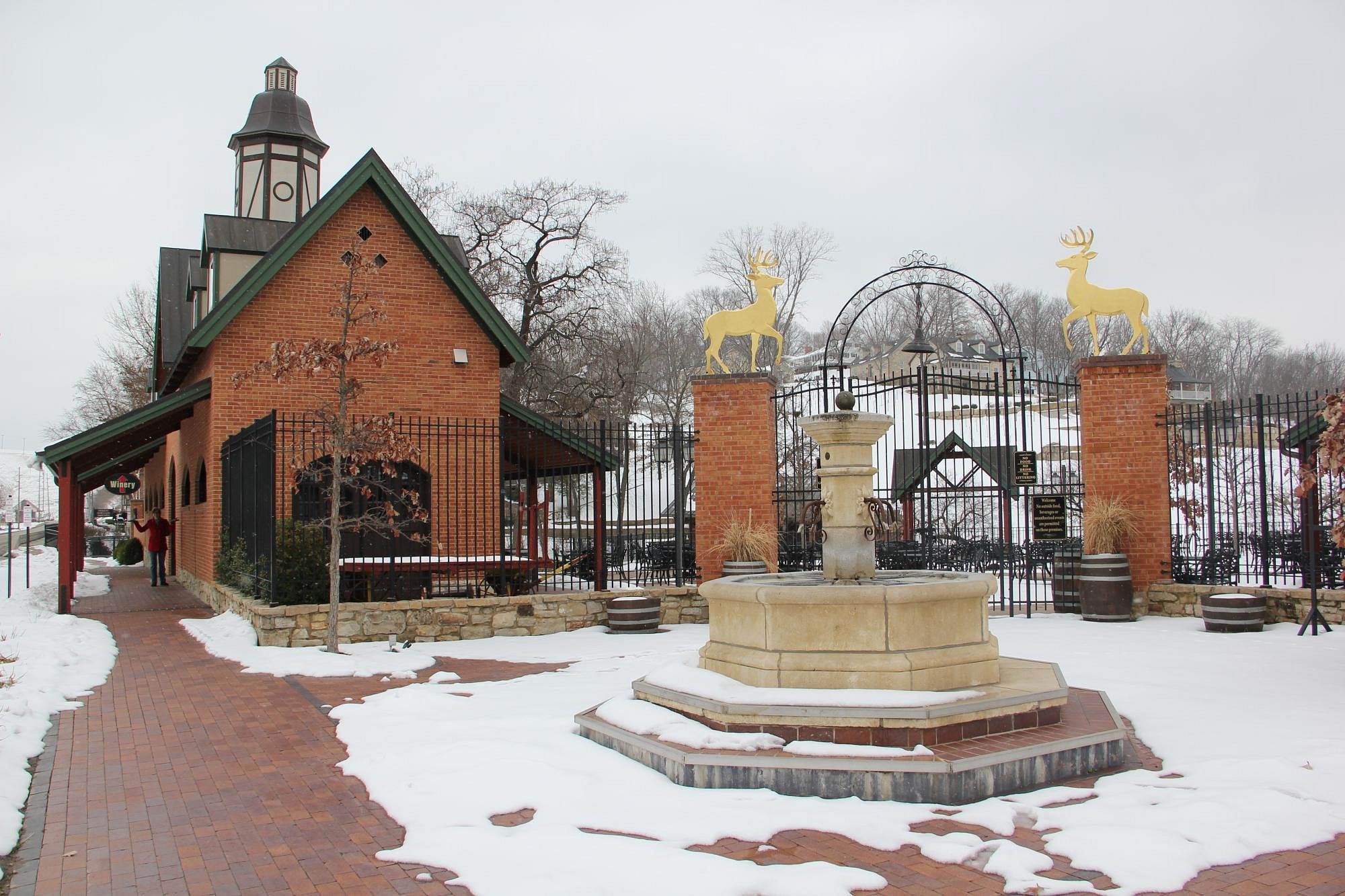 A brick building with a tower stands near a gated entrance topped with golden deer statues, evoking the charm of Unique American Towns. A stone fountain sits in front, snow blankets the ground, and bare trees and iron fences frame the scene.
