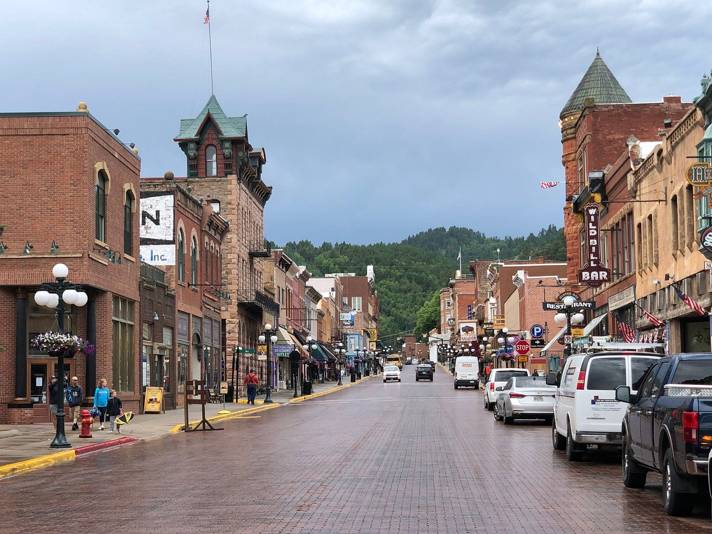 A view down a historic main street in one of those unique American towns, lined with brick buildings, shops, and bars. Cars are parked along both sides, with green forested hills rising in the background beneath a cloudy sky.
