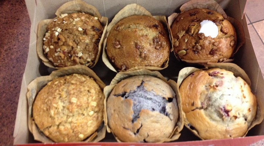 A box containing six assorted muffins, each with a different flavor and topping, arranged in two rows of three on a brown surface.