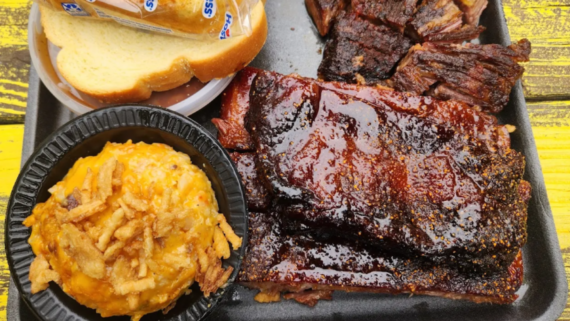 A tray from one of those hole-in-the-wall BBQ spots, stacked with barbecue glazed ribs, smoked brisket, white bread in plastic, and cheesy potato casserole topped with crispy onions&mdash;all set on a yellow wooden table.