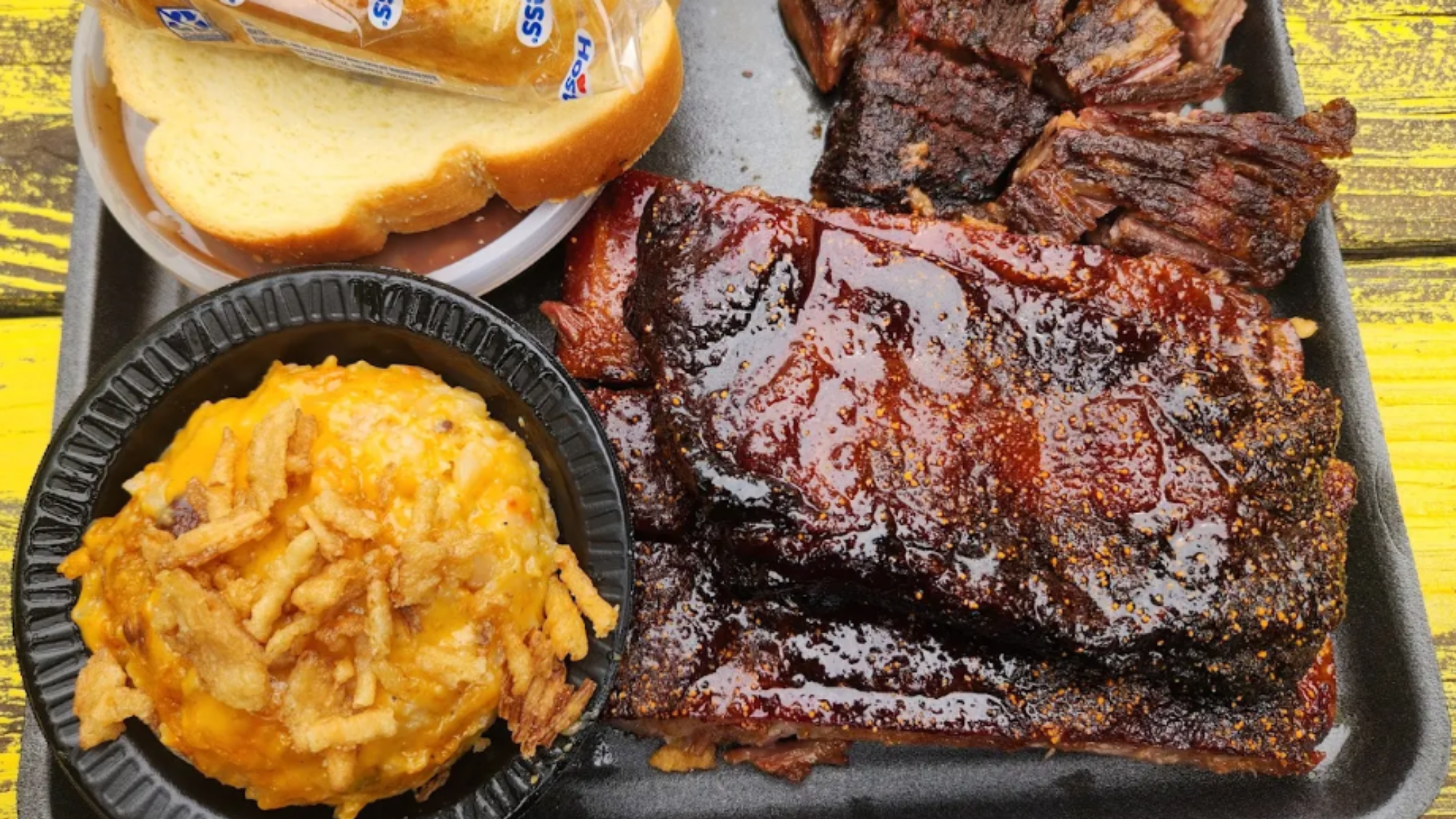 A tray from one of those hole-in-the-wall BBQ spots, stacked with barbecue glazed ribs, smoked brisket, white bread in plastic, and cheesy potato casserole topped with crispy onions—all set on a yellow wooden table.