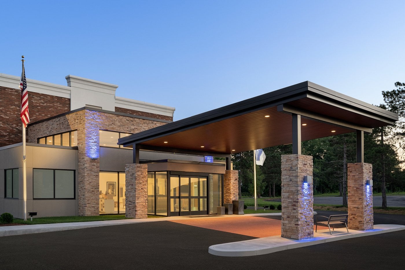 A modern Hotel Brands 2026 building with large glass doors, stone pillars, and a covered entrance canopy, illuminated by soft outdoor lights at dusk. An American flag is displayed near the entrance, with trees in the background.