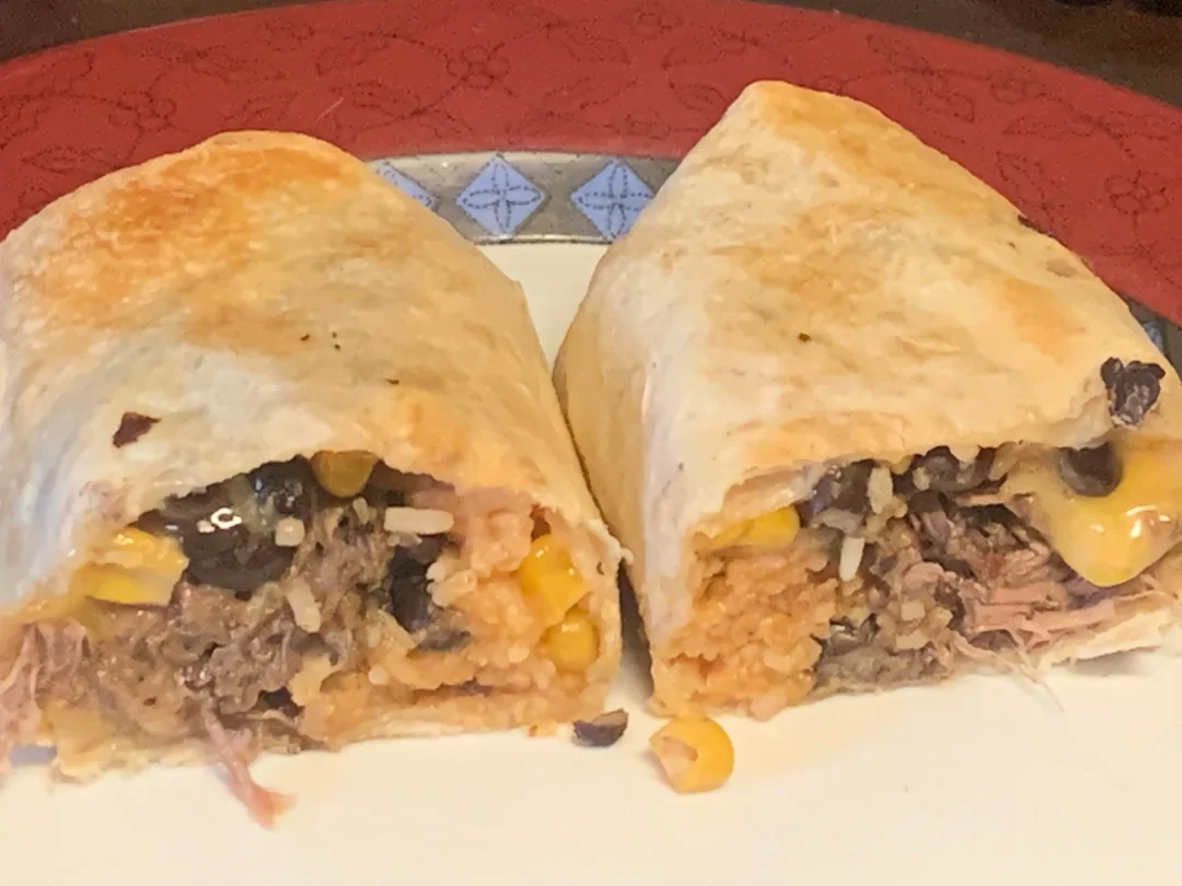 A burrito cut in half on a plate, showing a filling of shredded meat, rice, black beans, corn, and melted cheese, against a red tablecloth background.