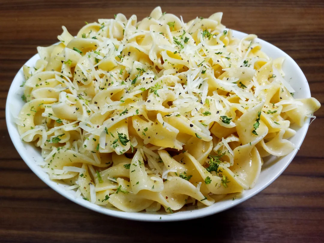 A white bowl filled with buttered egg noodles topped with grated cheese and chopped parsley, placed on a wooden surface.