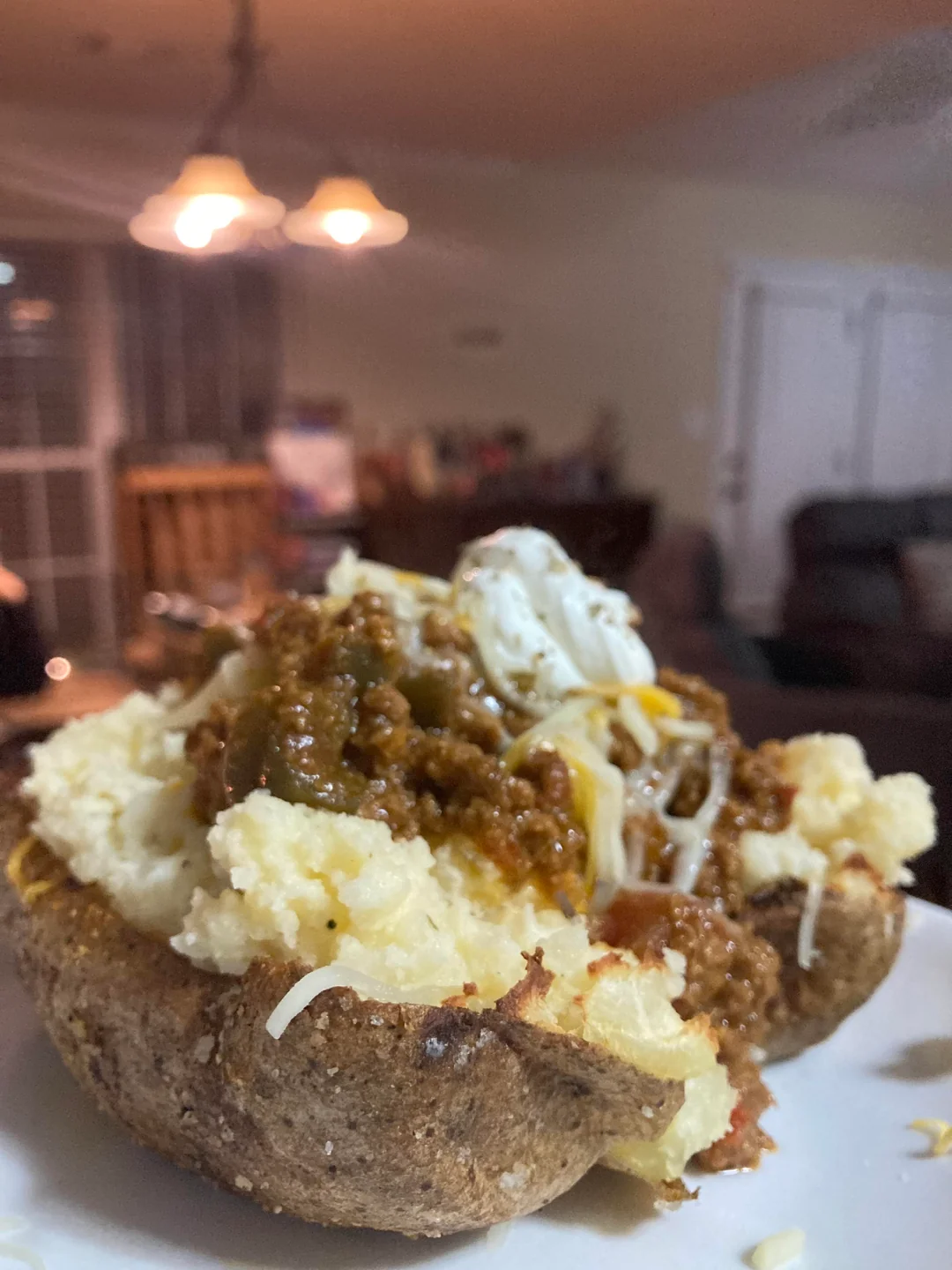 A baked potato topped with chili, shredded cheese, and a dollop of sour cream sits on a white plate, with a blurred living room in the background.