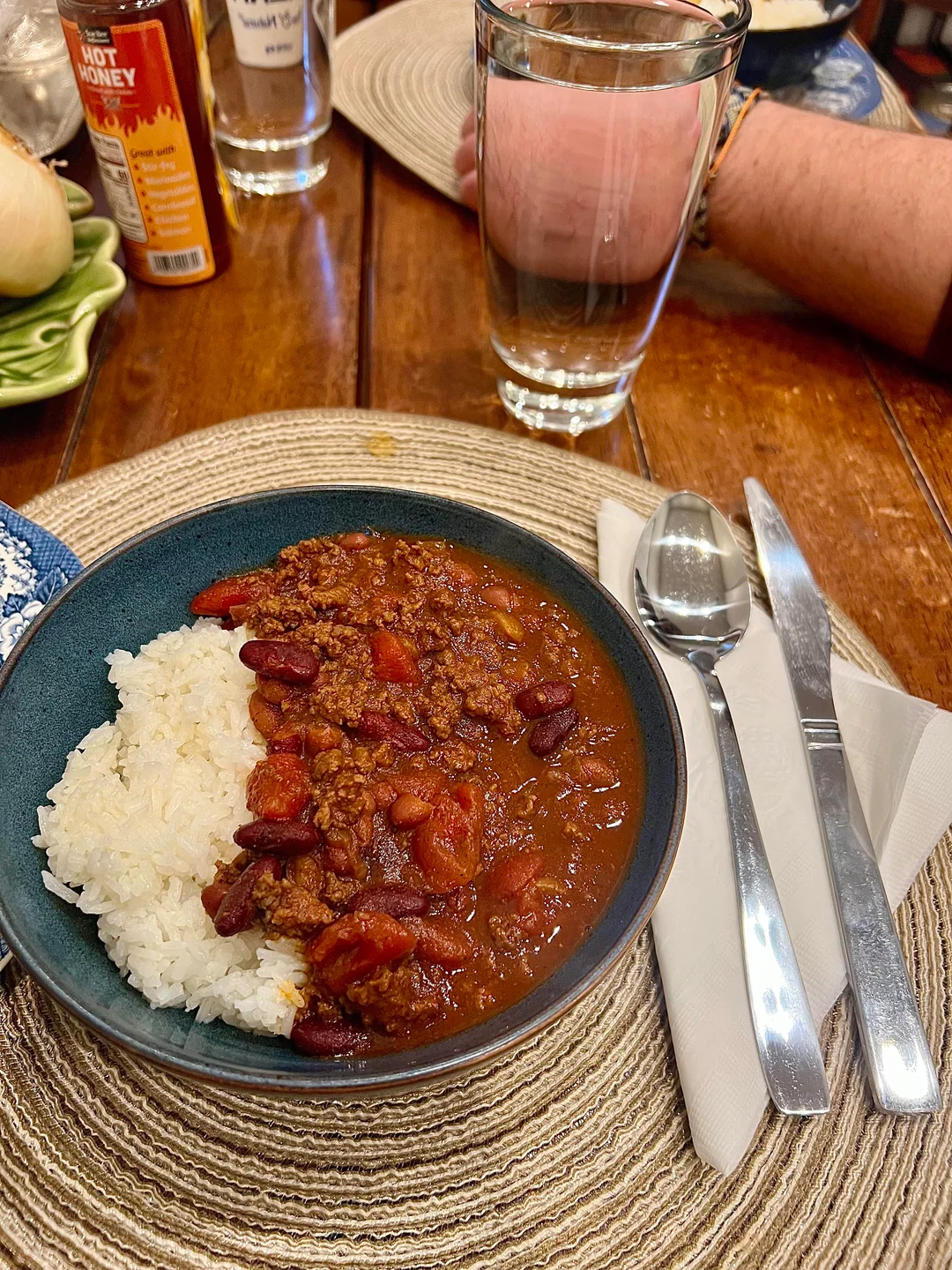 A bowl of chili with ground meat, red beans, and tomatoes served with white rice on a placemat. Next to the bowl are a fork, spoon, and knife on a napkin, with a glass of water and a person's arm in the background.