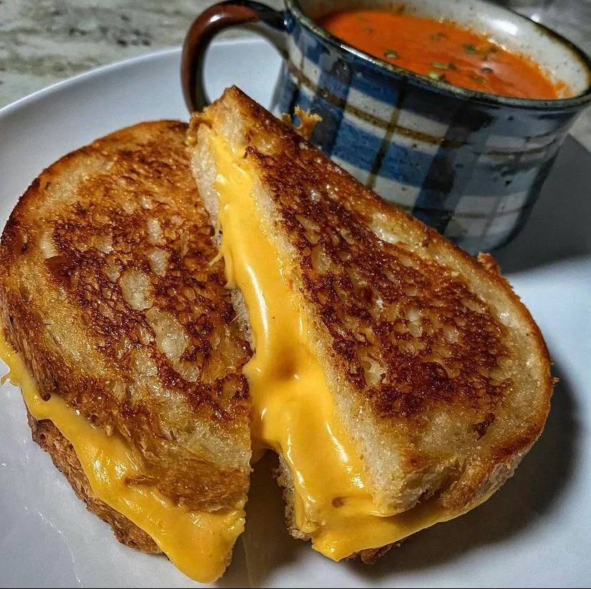 A golden, crispy grilled cheese sandwich cut in half with melted cheese oozing out, served on a white plate next to a mug of creamy tomato soup topped with herbs.