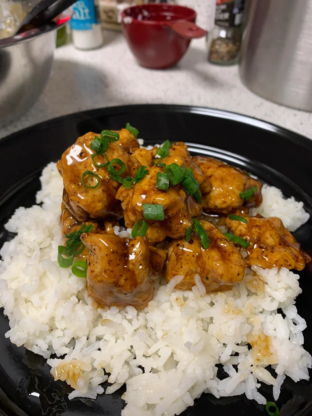 A plate of white rice topped with pieces of glazed chicken in a dark sauce, garnished with chopped green onions, served on a black dish with kitchen items in the background.